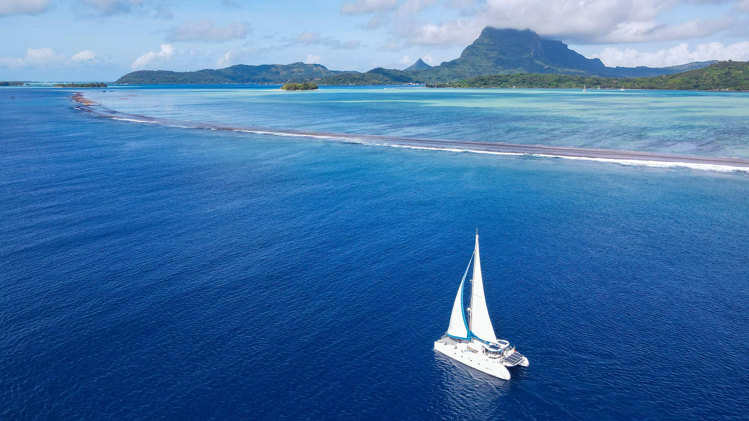 Catamaran sailboat with sails up sailing around Bora Bora on the edge of the reef with the lagoon and mountains beyond.