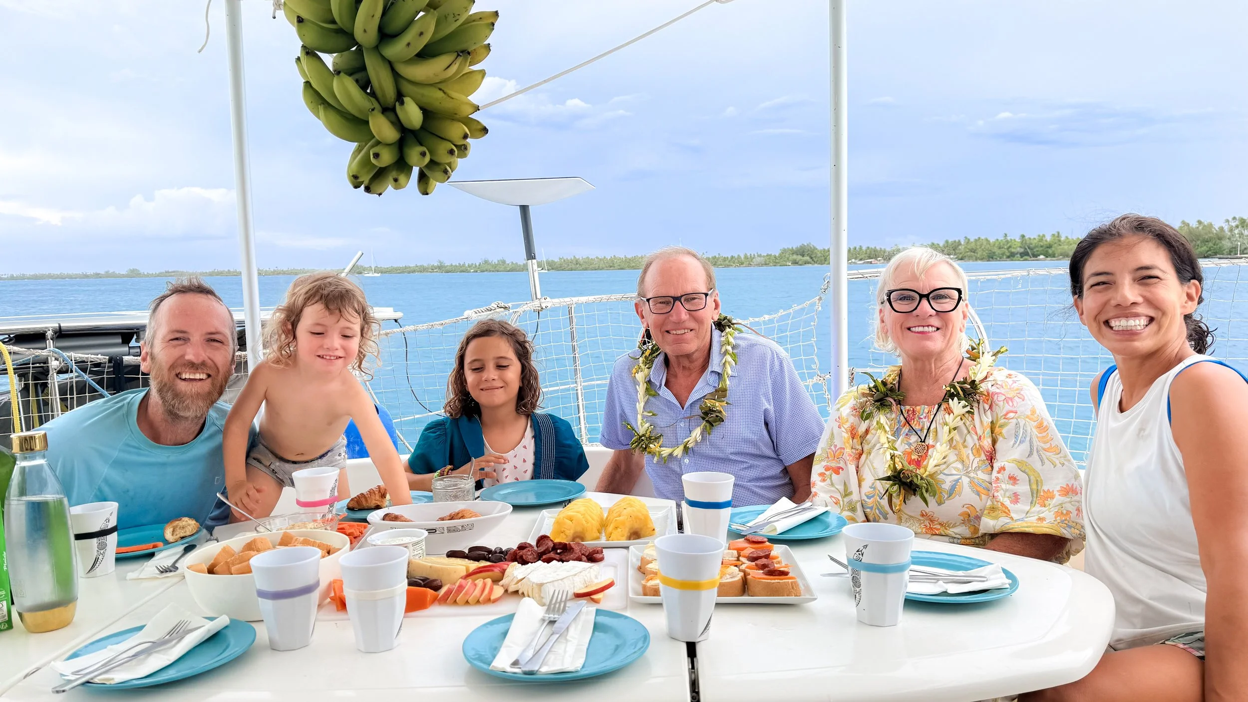 A group of people smiling and sitting around the table on a catamaran boat in a tropical setting about to eat a breakfast meal.
