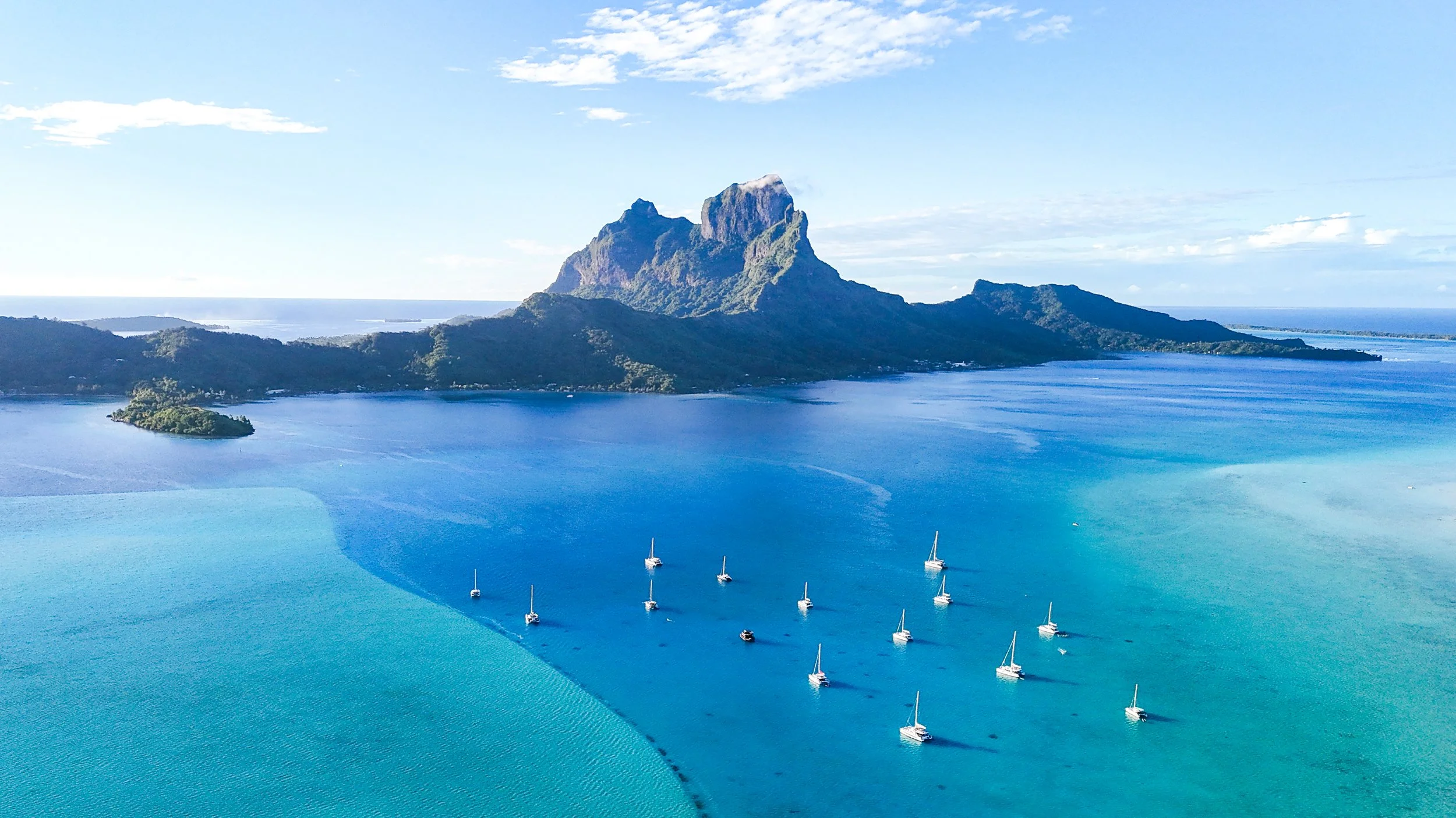 Aerial view of Bora Bora from turquoise and blue lagoon with white catamaran sailboats in foreground.