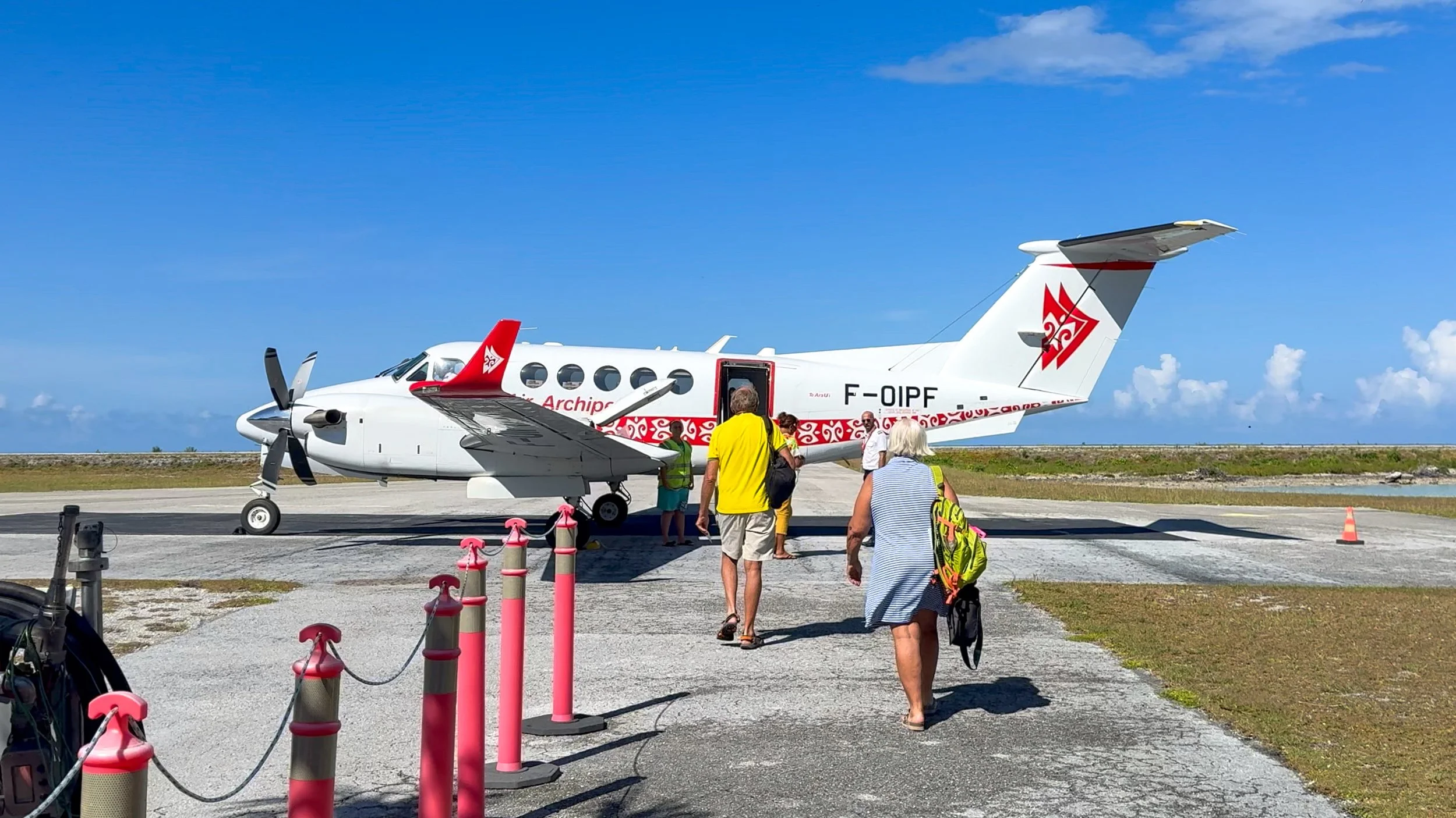 Small prop plane on runway at tropical airport with passengers walking outside to board.