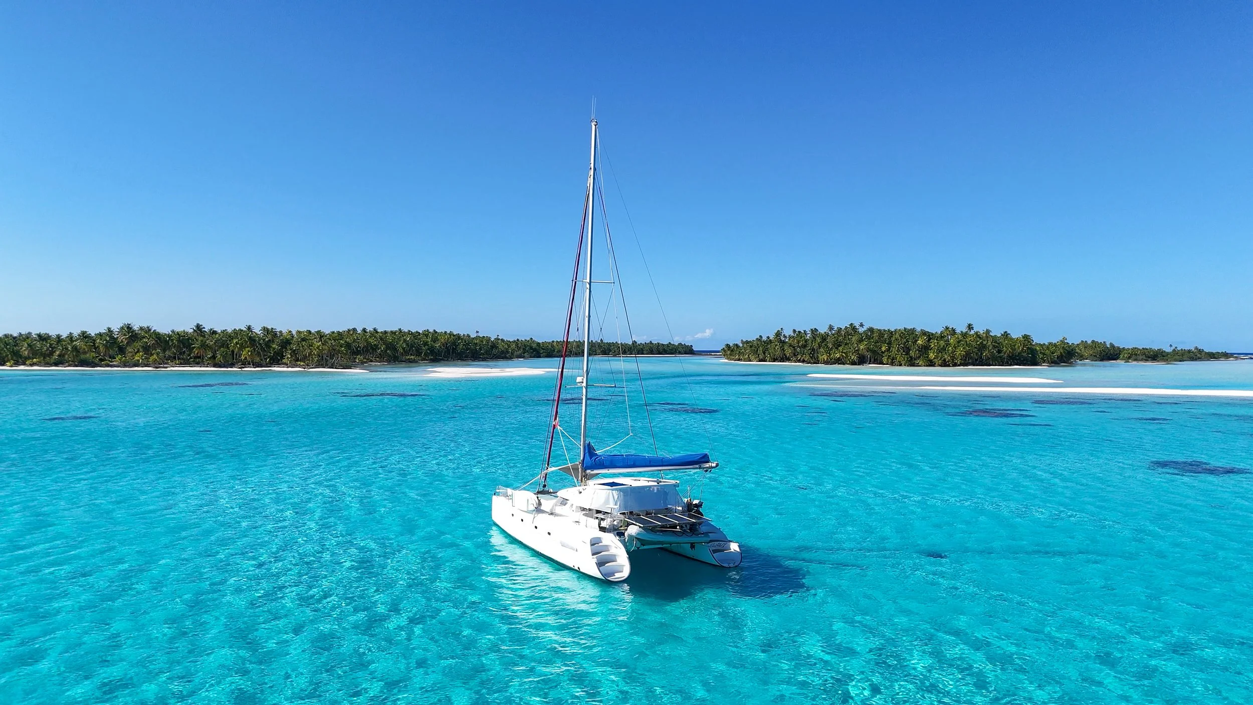 White catamaran sailboat floating in turquoise tropical water with palm trees on islands and white sand bars beyond.