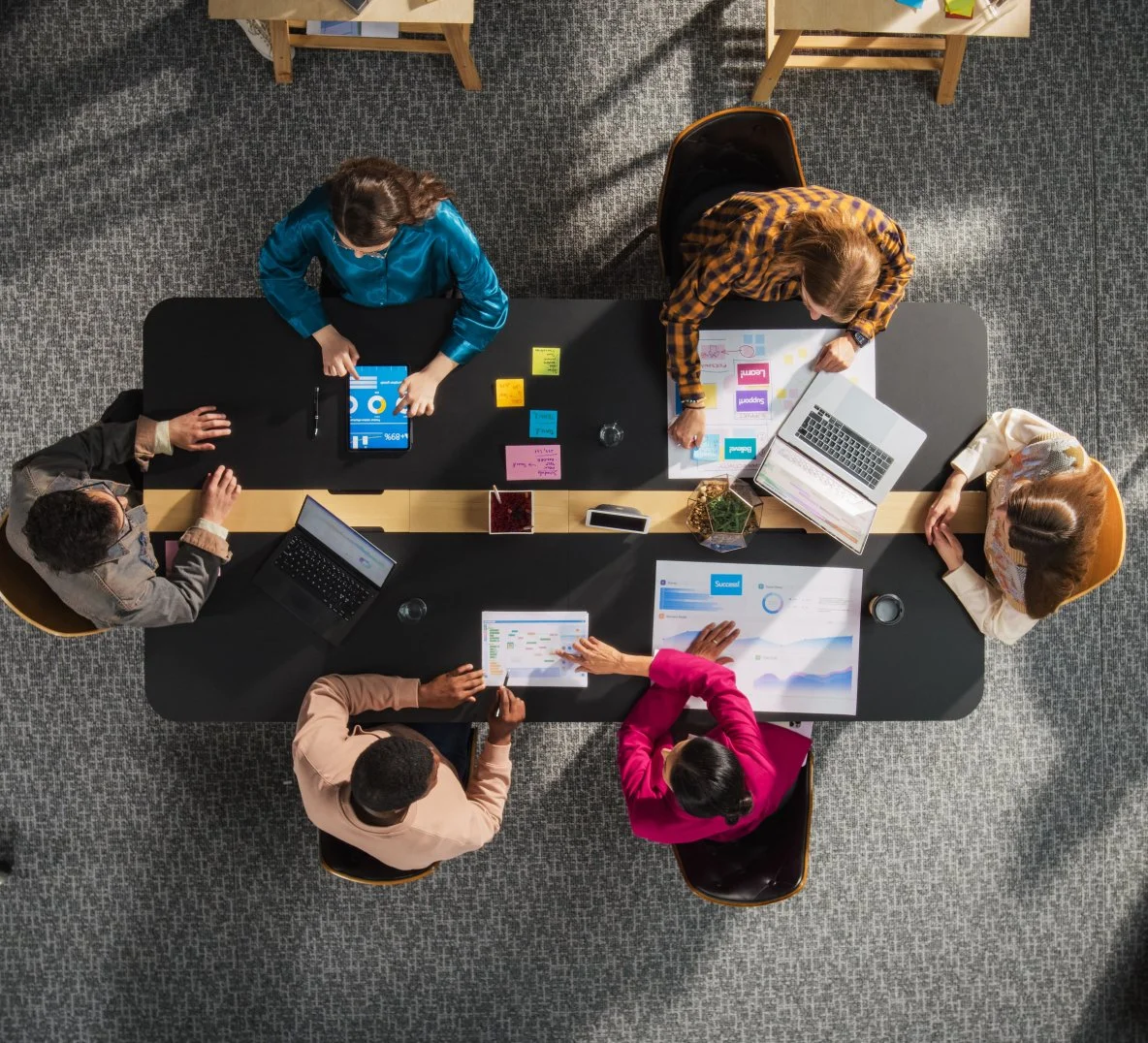 Overhead view of six diverse people working together around a black conference table with laptops, tablets, and colorful sticky notes, in a modern office.