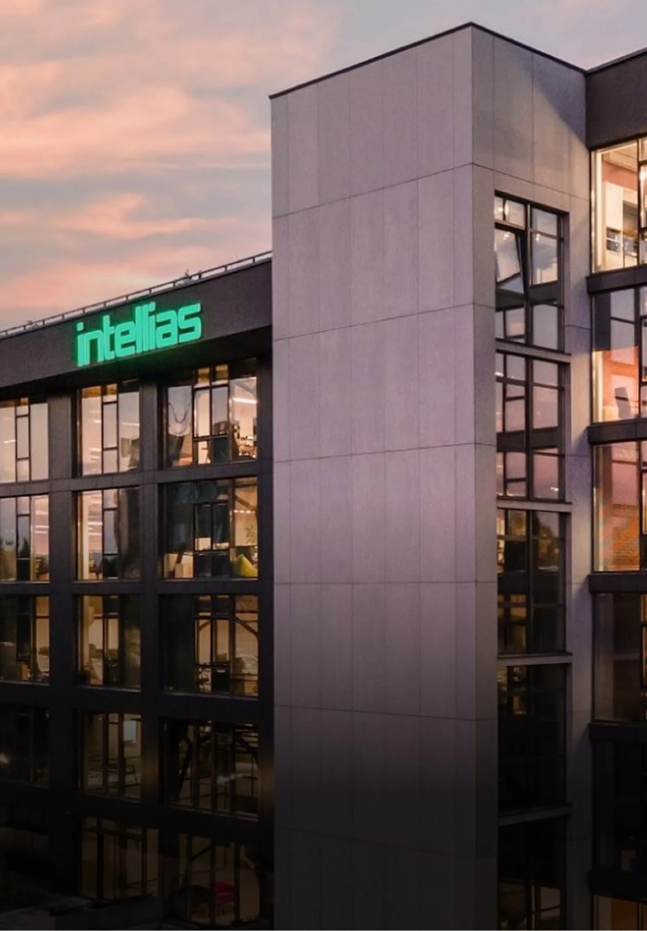 Exterior view of a modern glass office building with a green illuminated sign reading 'Intellias' at sunset.
