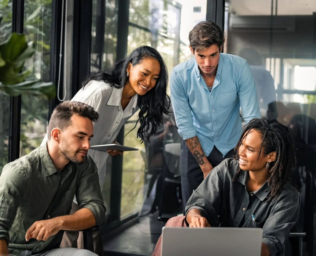 A diverse group of four young professionals collaborating, with two men and two women, gathered around a laptop in a modern office with large windows.