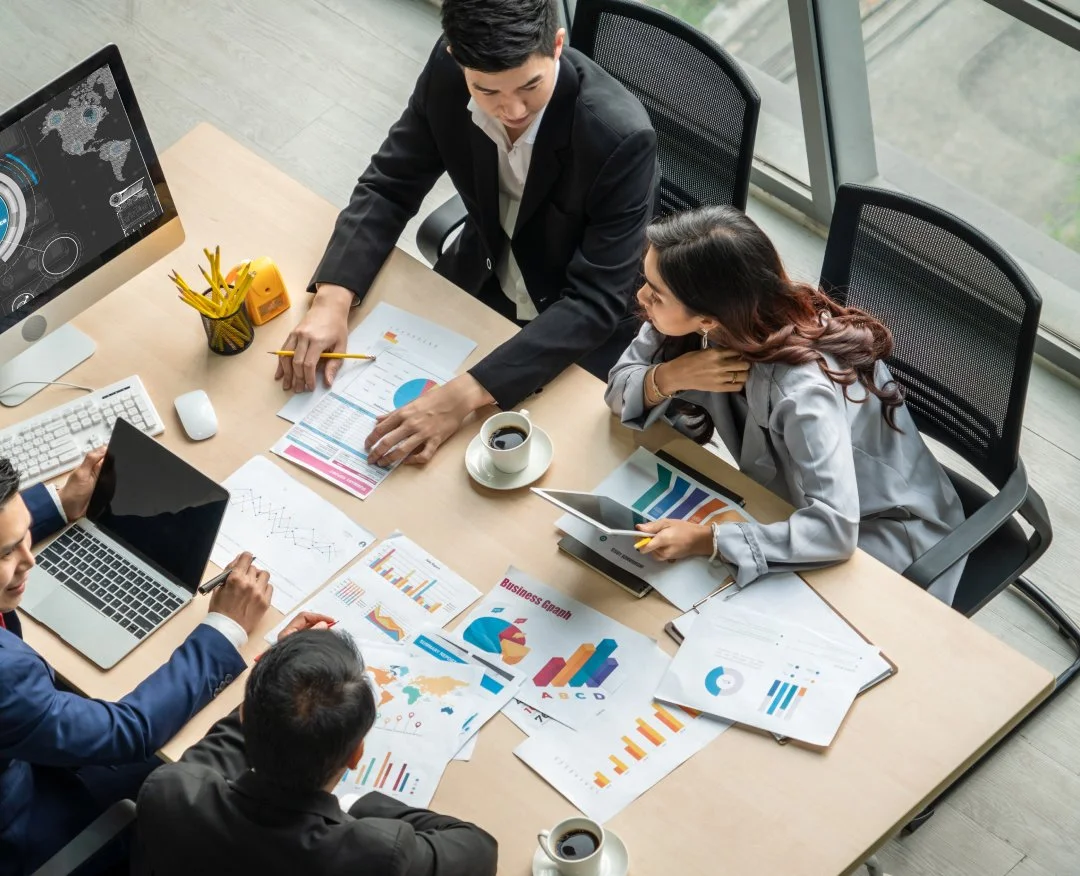 A group of five people in business attire having a meeting in a modern office with large windows. They are discussing various charts and graphs related to business data, with laptops, tablets, and coffee cups on the table.