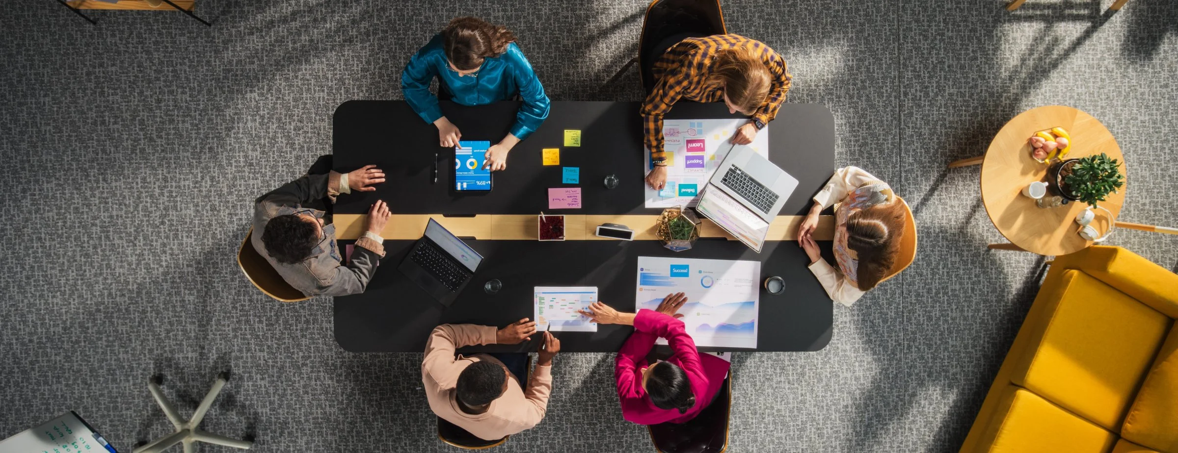 Top-down view of six professionals sitting around a black conference table, working on laptops, tablets, and printed charts, with sticky notes and office supplies on the table, in a well-lit modern office.