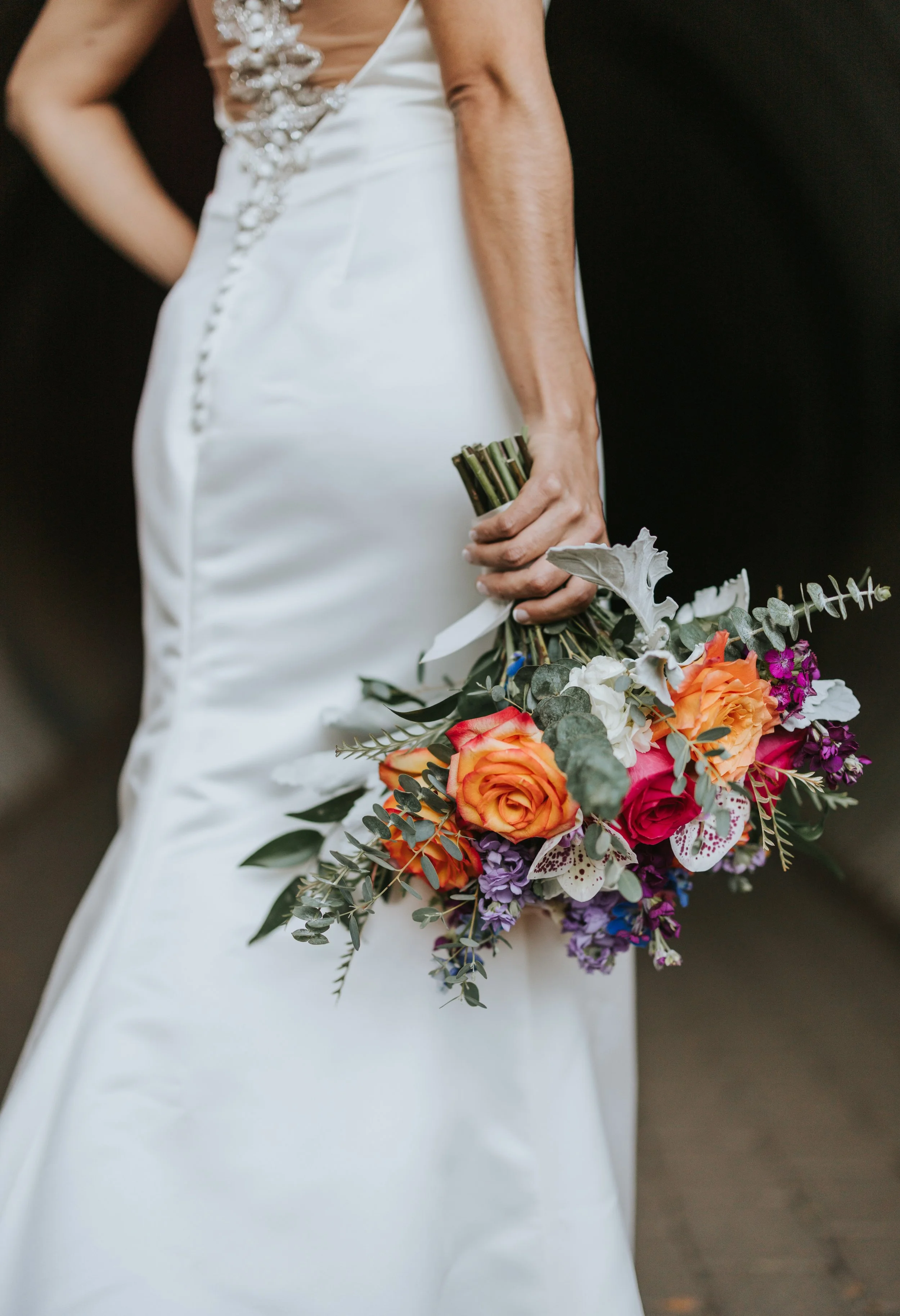 Close-up of a woman in a white wedding dress holding a colorful bouquet of flowers against a dark background.