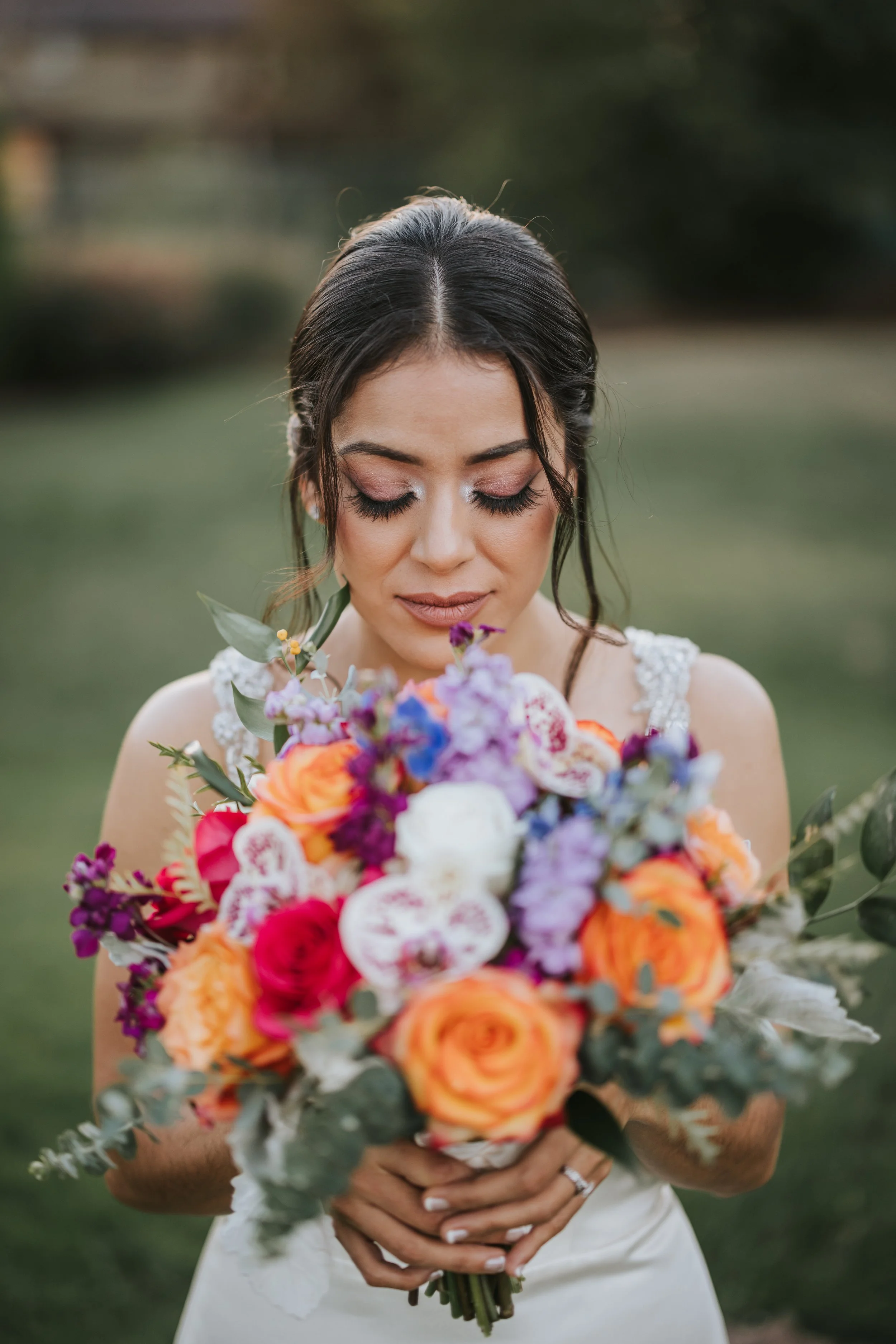 A bride with dark hair, closed eyes, and light makeup, holding a large colorful bouquet of flowers, standing outdoors in a garden or park setting.