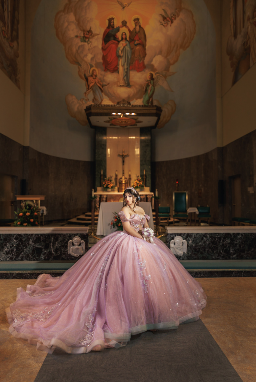 A young woman in a pink ball gown with embellishments, wearing a tiara and holding a bouquet, standing inside a church or chapel with religious artwork, altar, and floral decorations.