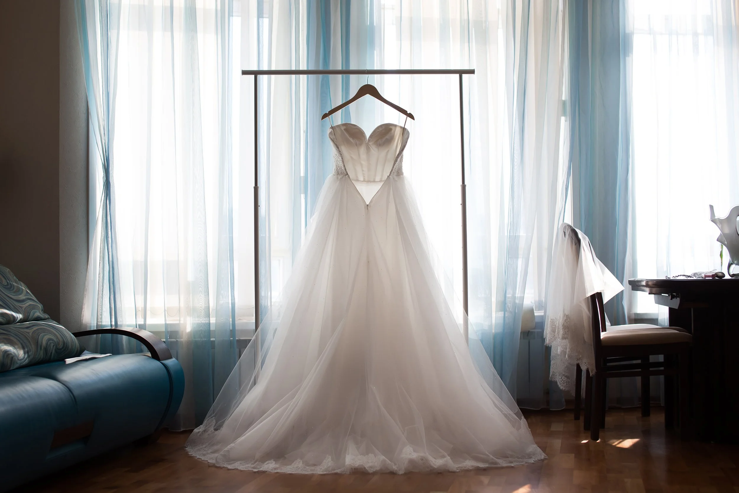 Wedding dress hanging on a wooden hanger in front of a window with blue and white curtains.