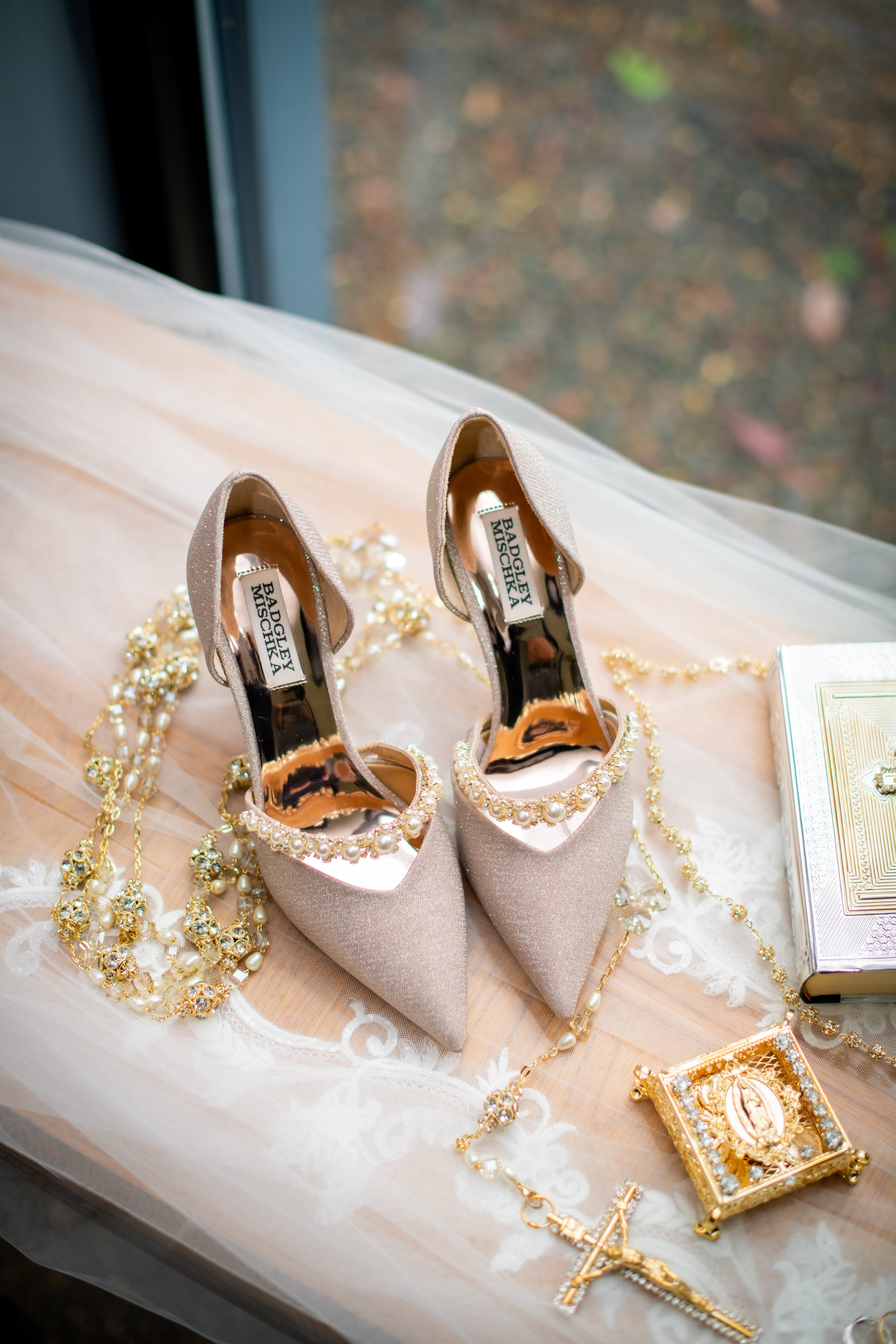 A pair of beige glitter high heel shoes decorated with pearl and rhinestone embellishments, placed on a lace and tulle fabric. Surrounding the shoes are bridal jewelry, including a long pearl necklace, a gold cross necklace, a framed religious icon, 