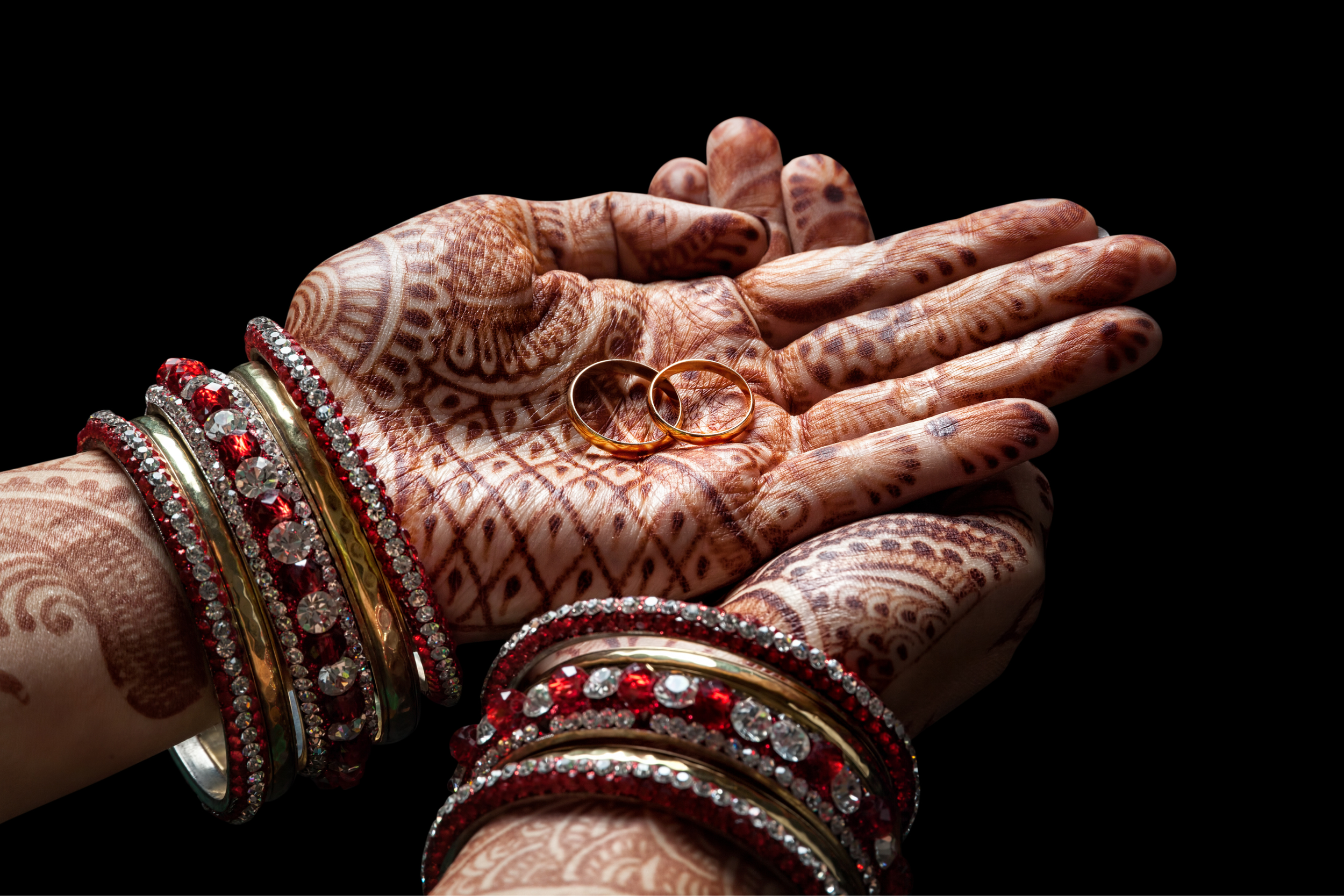Hand with intricate henna designs holding two gold wedding rings, decorated with red and silver bangles against a black background.