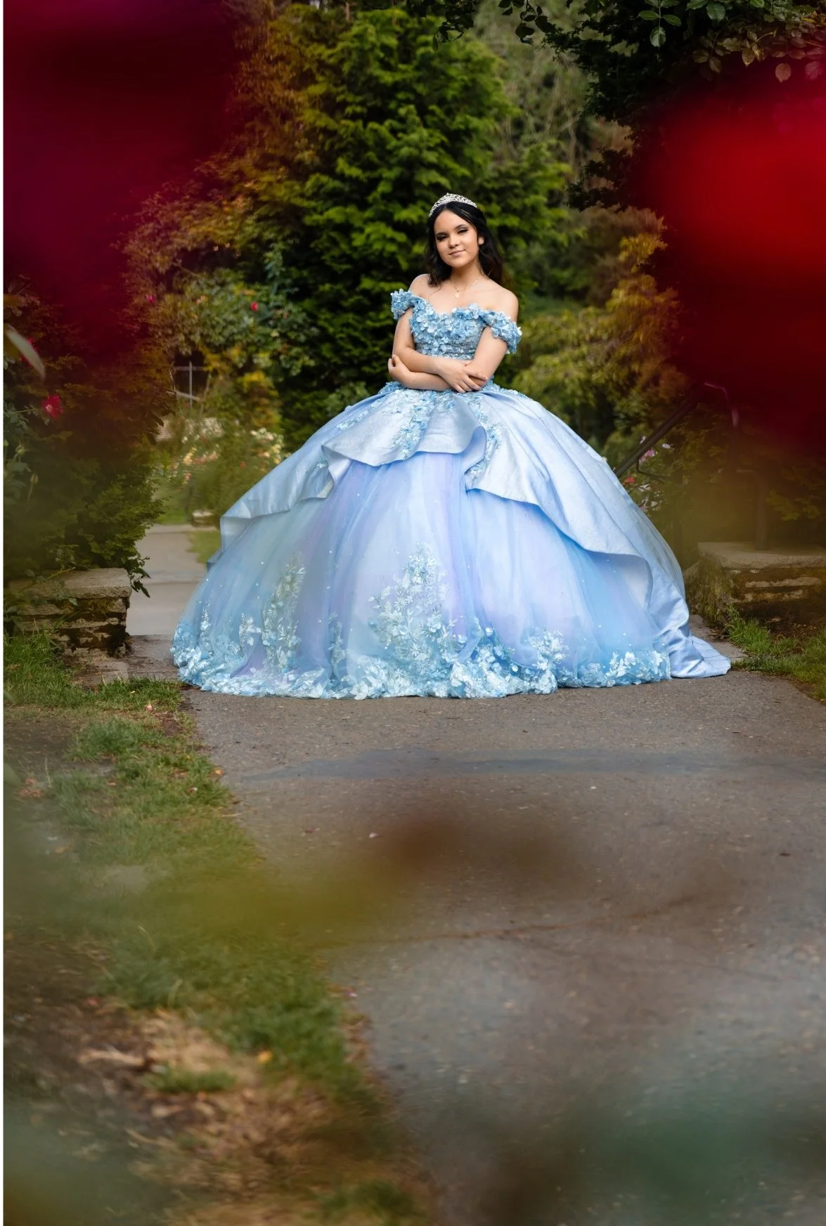 Young woman in a light blue princess dress standing on a paved path surrounded by greenery and trees.