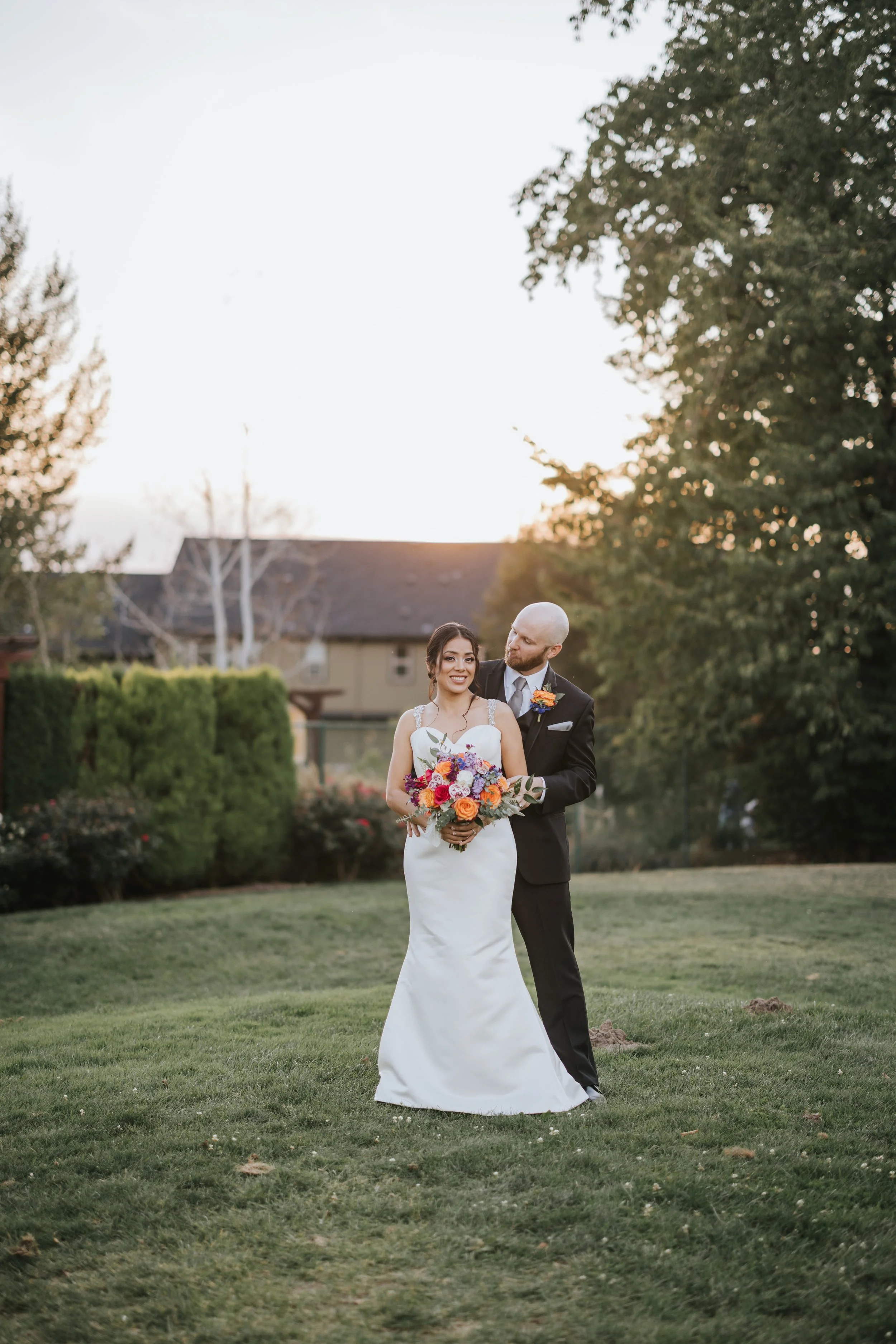 Bride in a white wedding gown holding a colorful bouquet, standing next to groom in a black suit with boutonniere, outdoors on grass during sunset