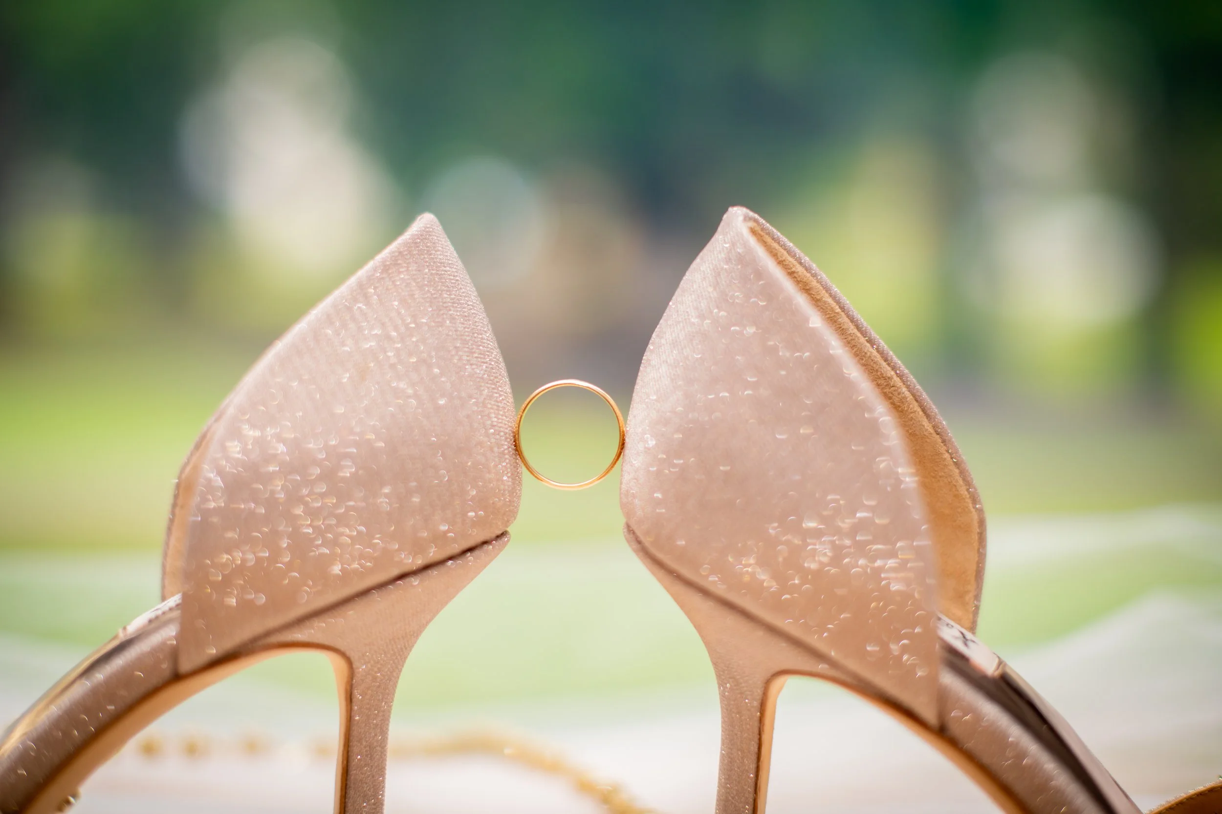 A pair of glittery beige high-heeled shoes facing each other with a wedding ring hanging between the heels, against a blurred outdoor background.
