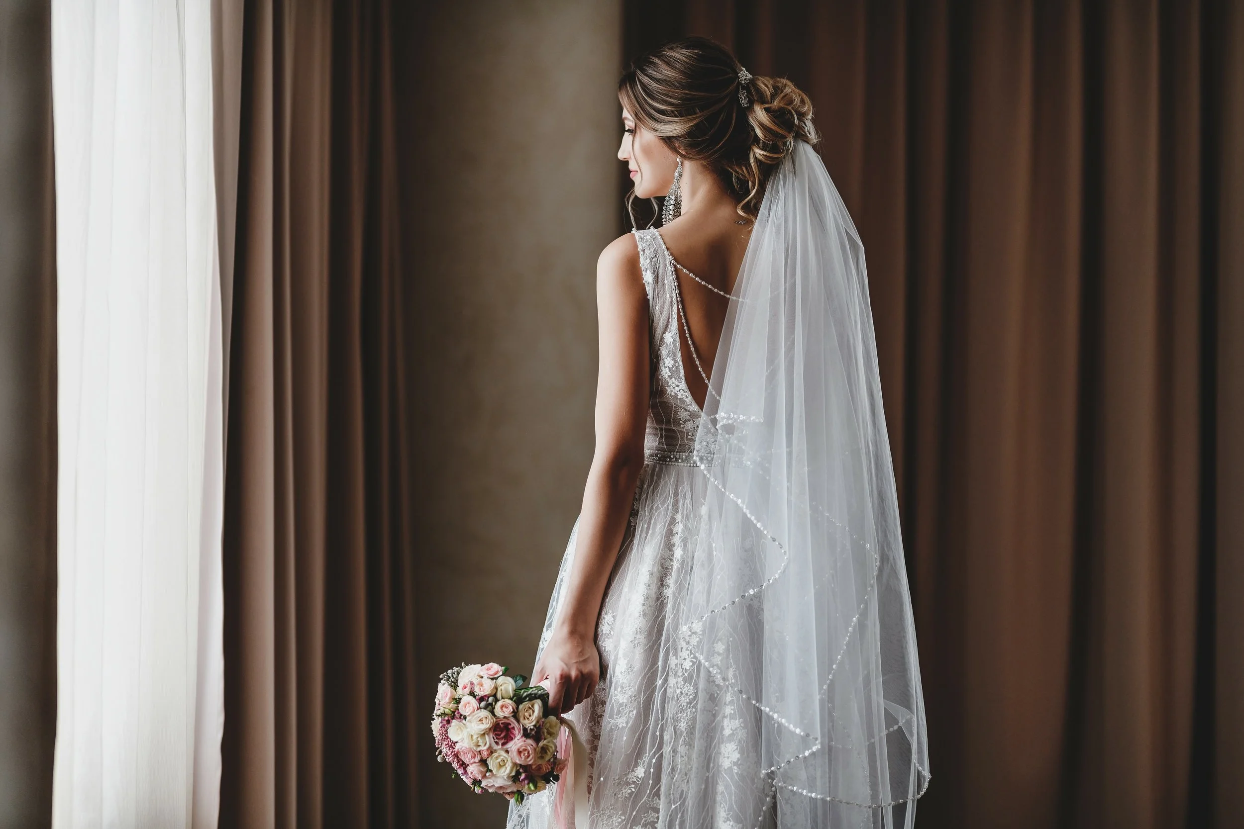A bride in a lace wedding dress holding a bouquet, standing by a window with beige curtains.