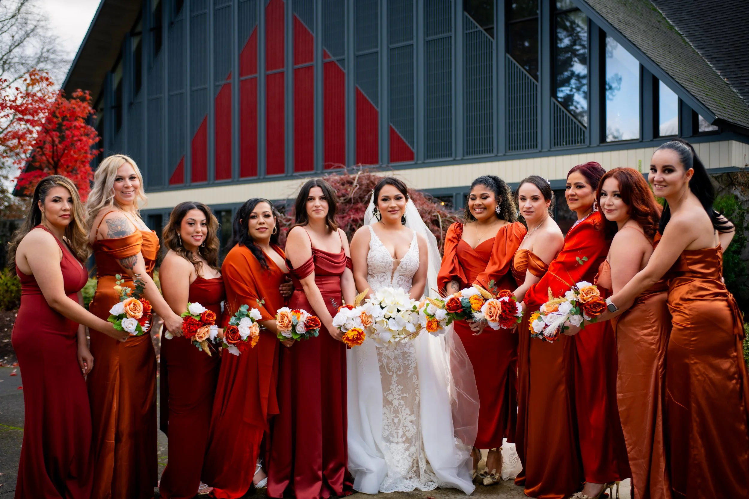 Bride in white wedding dress surrounded by ten bridesmaids in red and orange dresses holding bouquets, standing outdoors in front of a modern building with red and black accents and autumn trees.