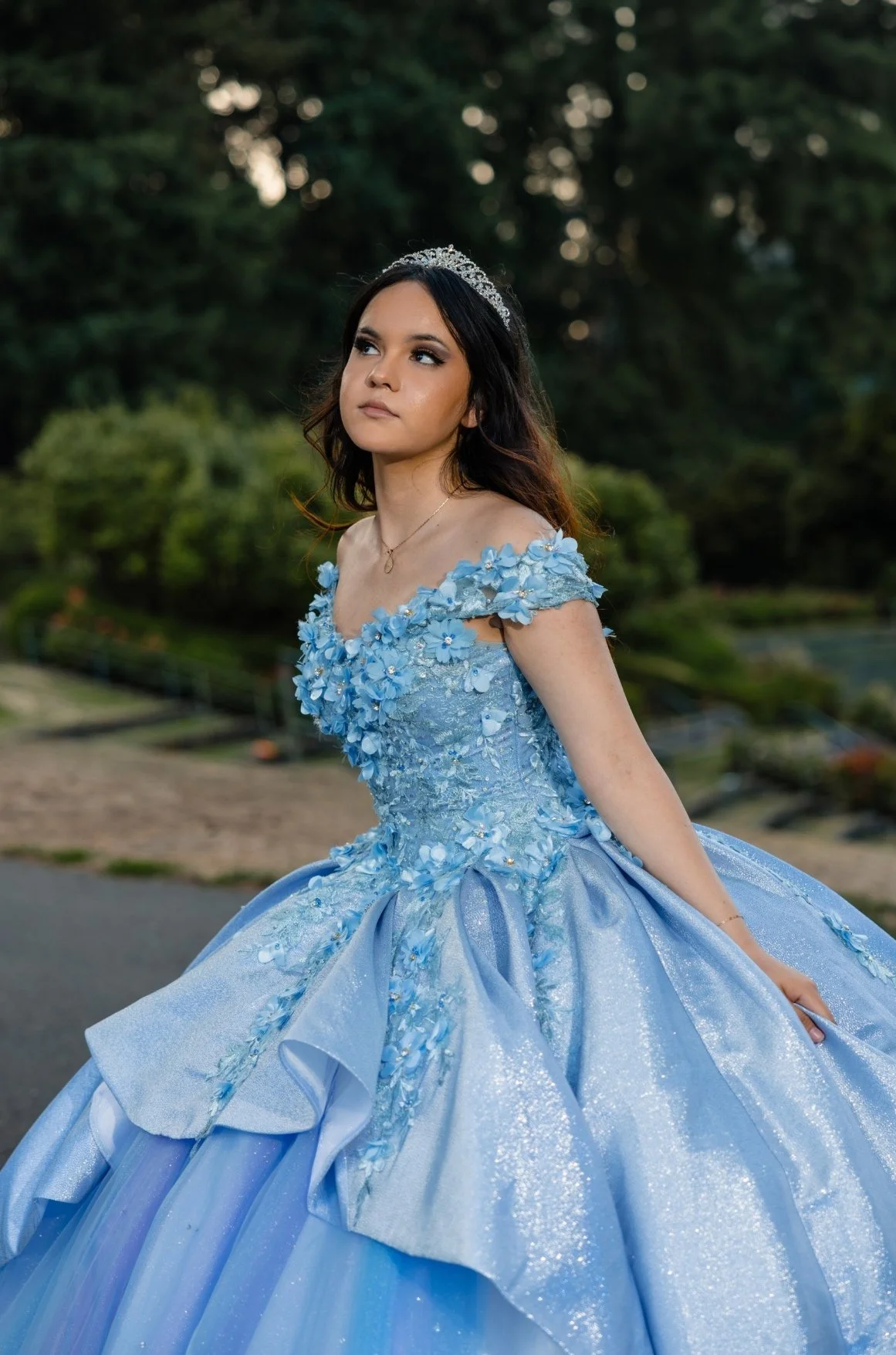A young woman wearing a blue ball gown with floral embellishments, a tiara, and a delicate necklace standing outdoors with a blurred background of trees and greenery.