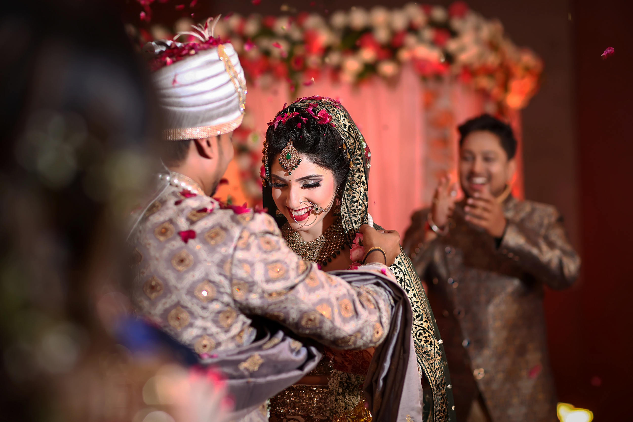 Indian bride and groom in traditional wedding attire exchanging garlands, with a smiling groomsman in the background, during a wedding ceremony decorated with pink flowers.