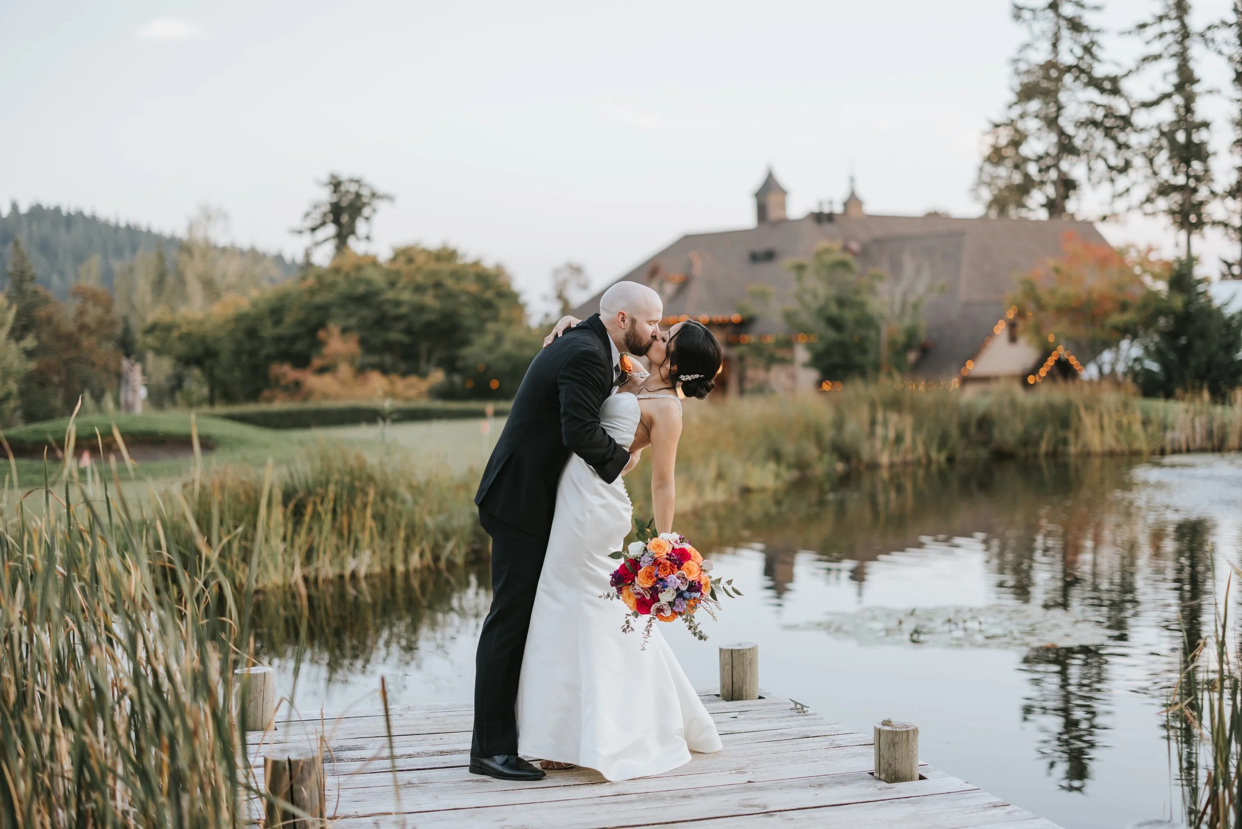 A bride and groom sharing a kiss on a wooden dock by a lake, with a large house and trees in the background during sunset.
