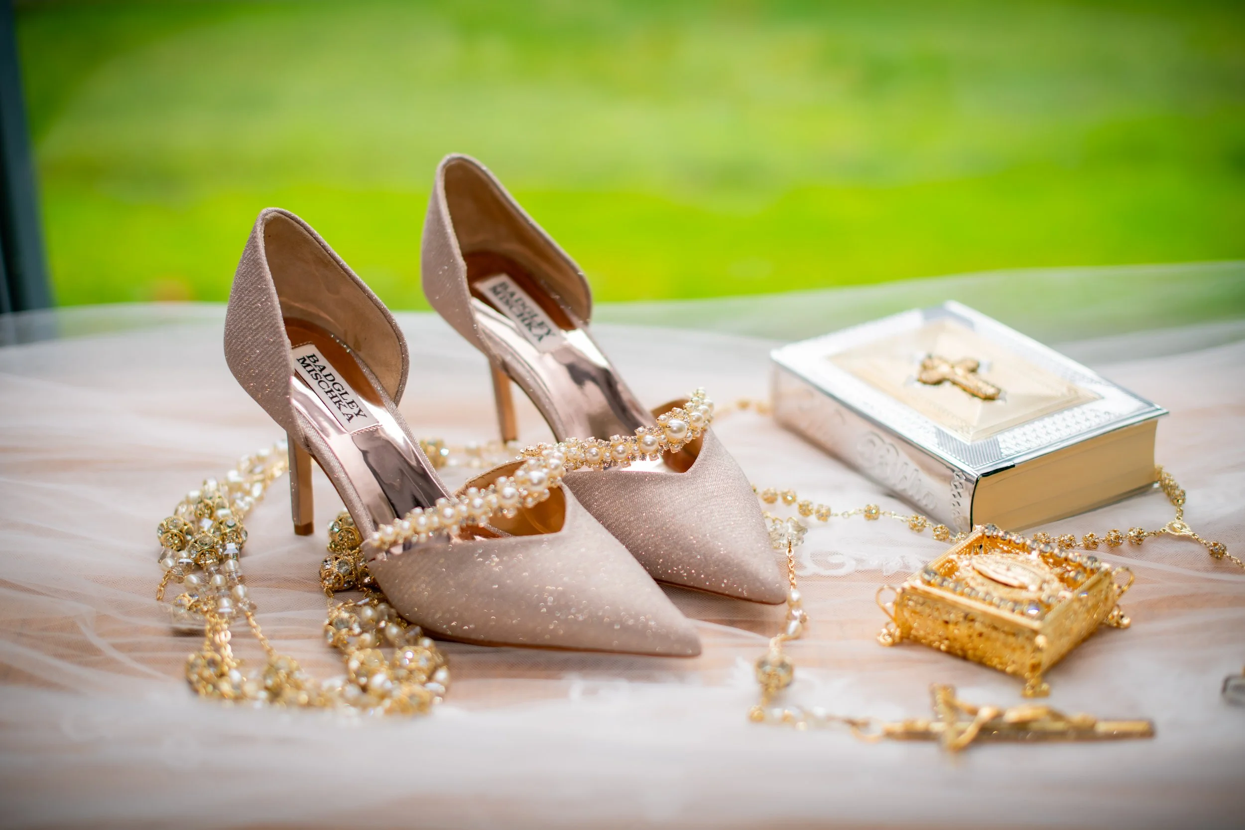 Pair of glittery high-heeled shoes surrounded by pearl necklaces, a decorative jewelry box, and a wedding or religious book on a soft cloth with a blurred green background.