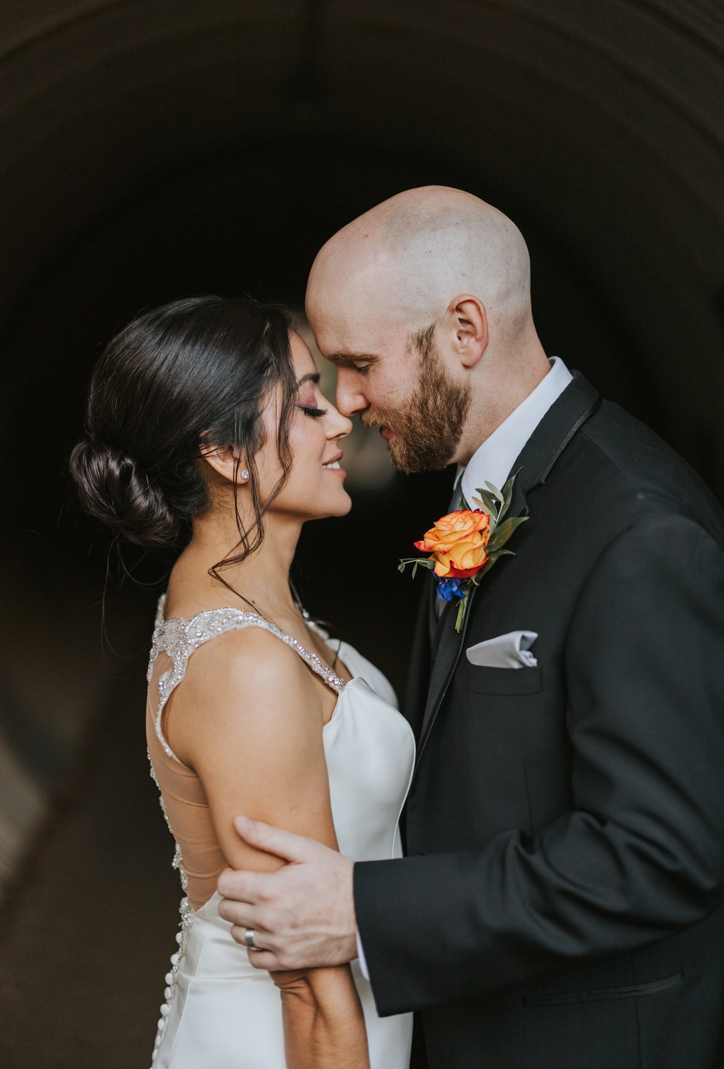 A newlywed couple is close together, touching foreheads and smiling, standing in a dark tunnel. The woman has dark hair styled in an elegant updo and is wearing a white wedding dress with beaded shoulder details. The man has a shaved head, beard, and