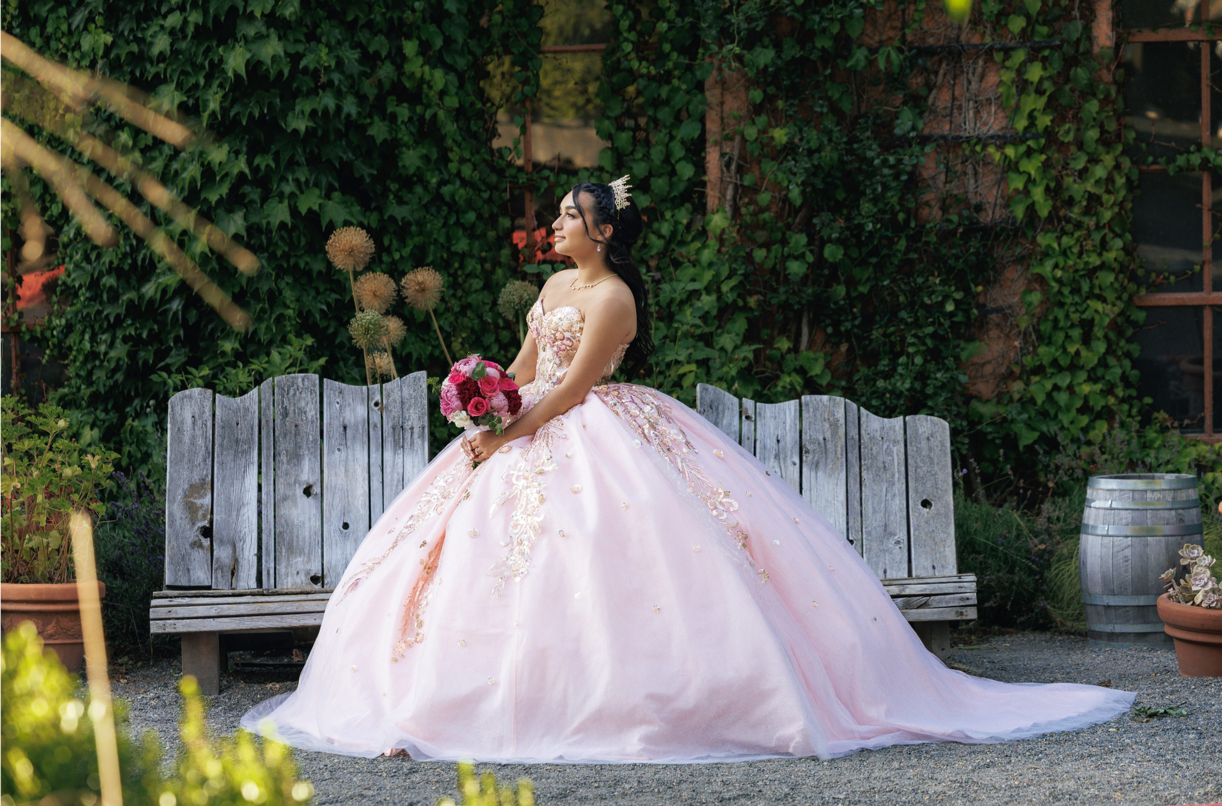 A woman in a pink, embellished ball gown and tiara holding a bouquet, standing outdoors in front of a wooden fence and green ivy-covered wall.