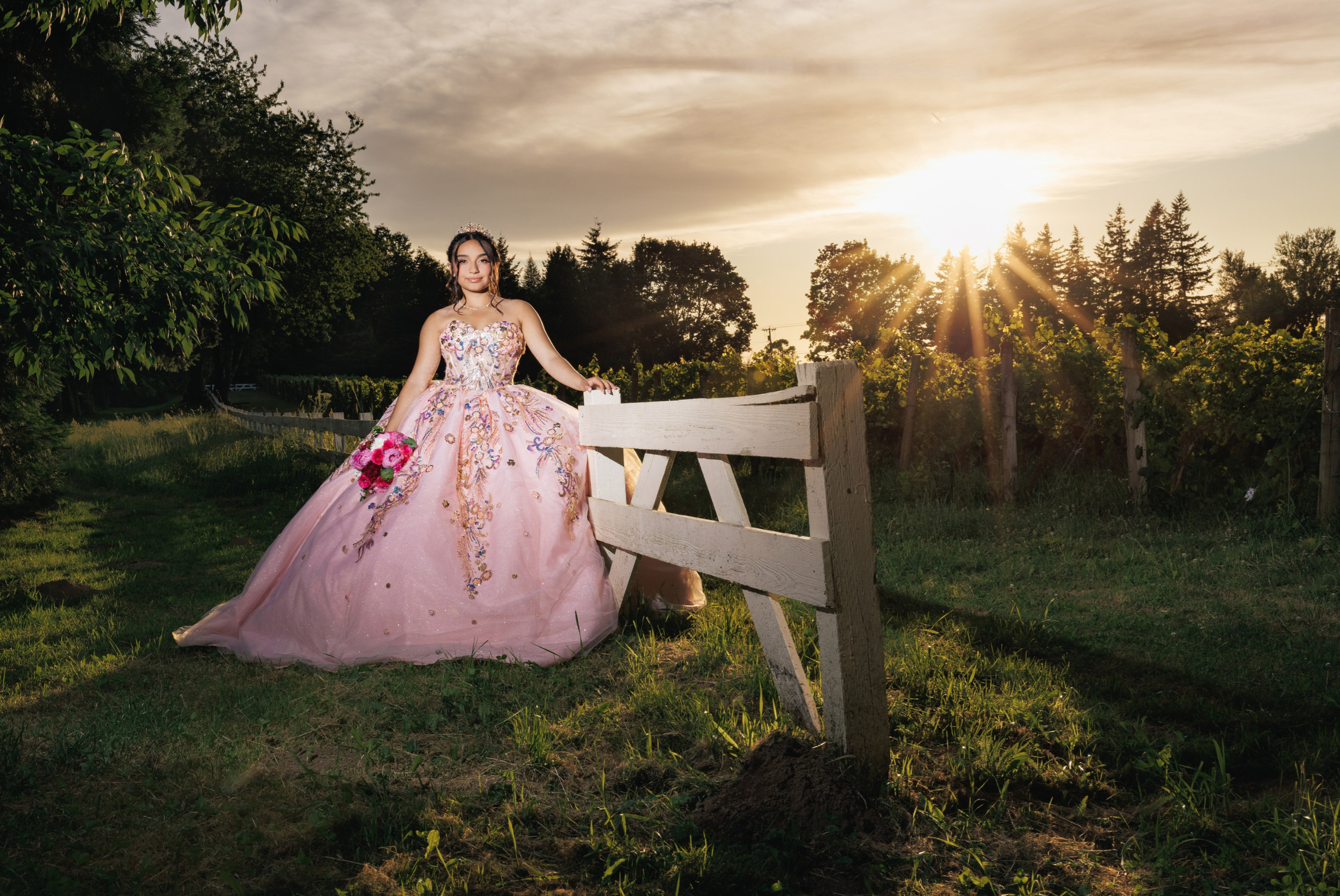 Young woman in a decorated pink ball gown holding a bouquet of pink flowers outdoors at sunset.