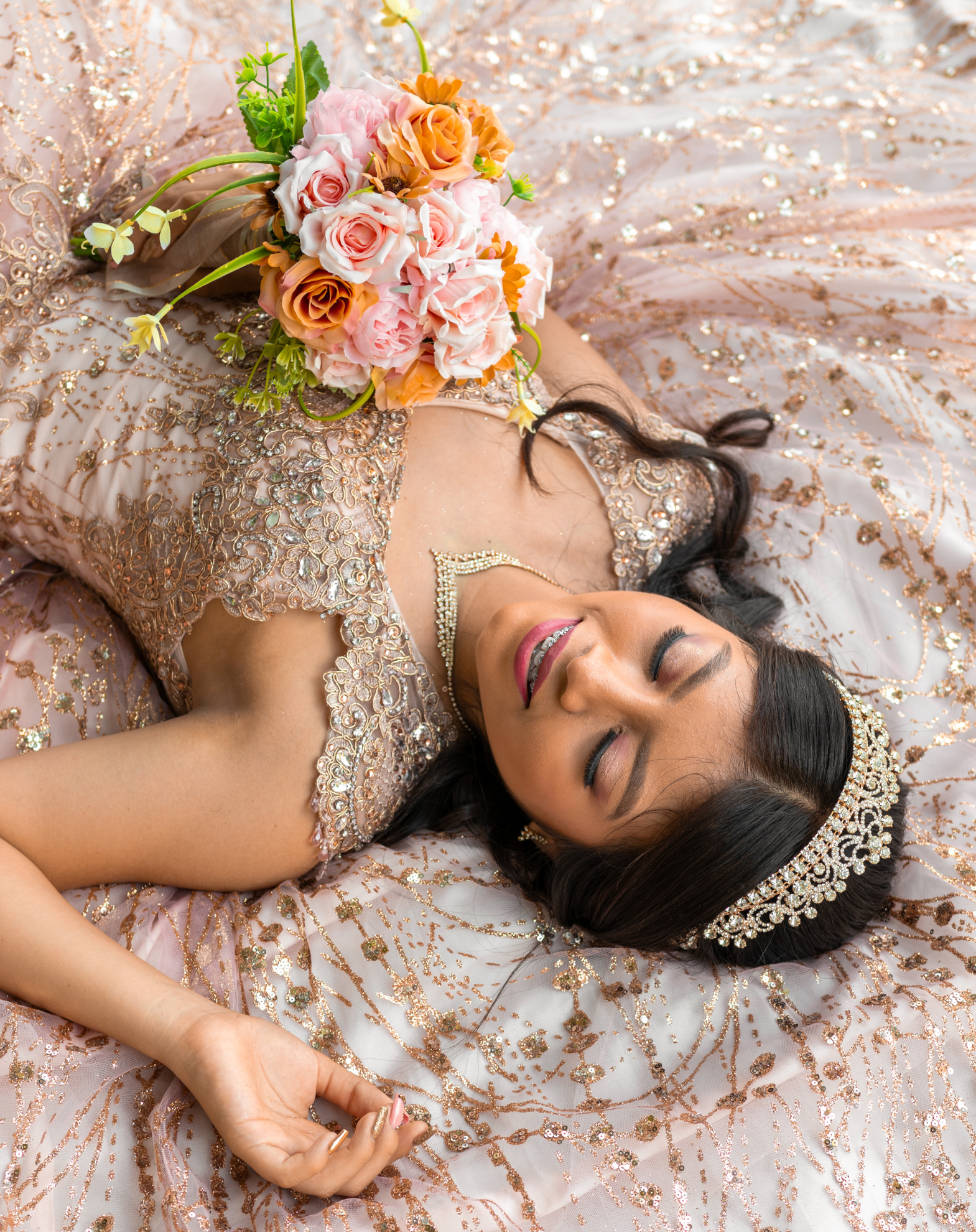 A bride lying on a bejeweled pink and gold dress, holding a pink and orange floral bouquet, wearing a tiara and jewelry, smiling with eyes closed.