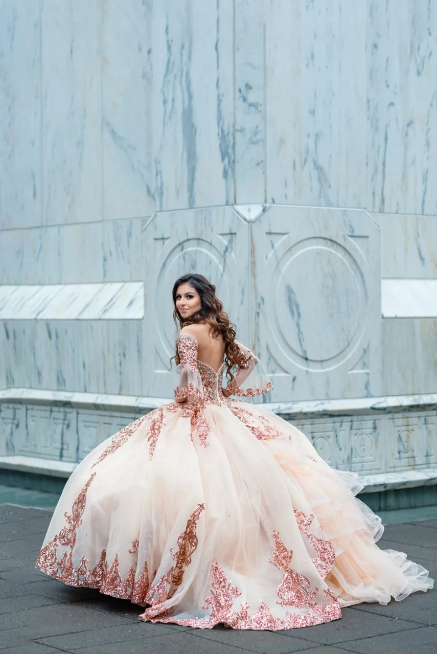 A woman in an elegant, off-the-shoulder, pink and gold ball gown with intricate embroidery, standing outdoors in front of a marble wall.