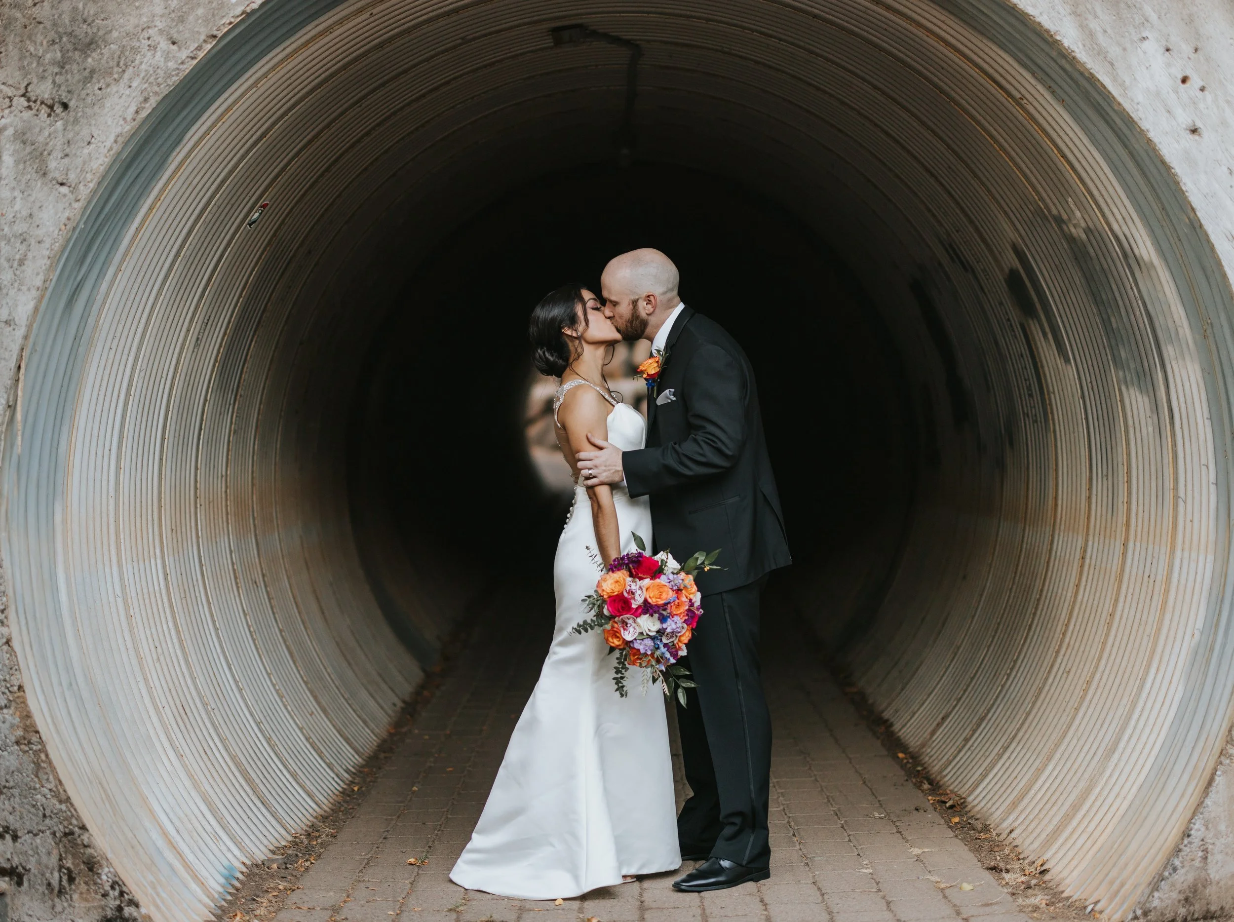 A bride and groom share a kiss inside a large, circular metal tunnel, with the bride holding a colorful bouquet of flowers.