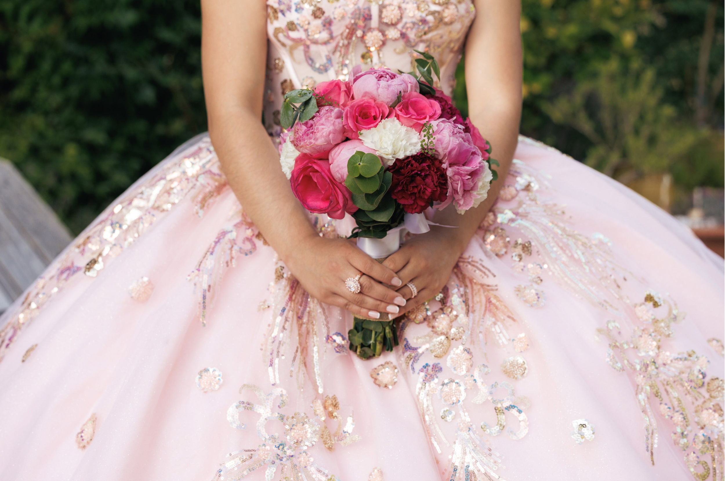 A woman in an elegant pink gown with intricate embroidery and beadwork, holding a bouquet of pink and white flowers with greenery, outdoors in a garden setting.