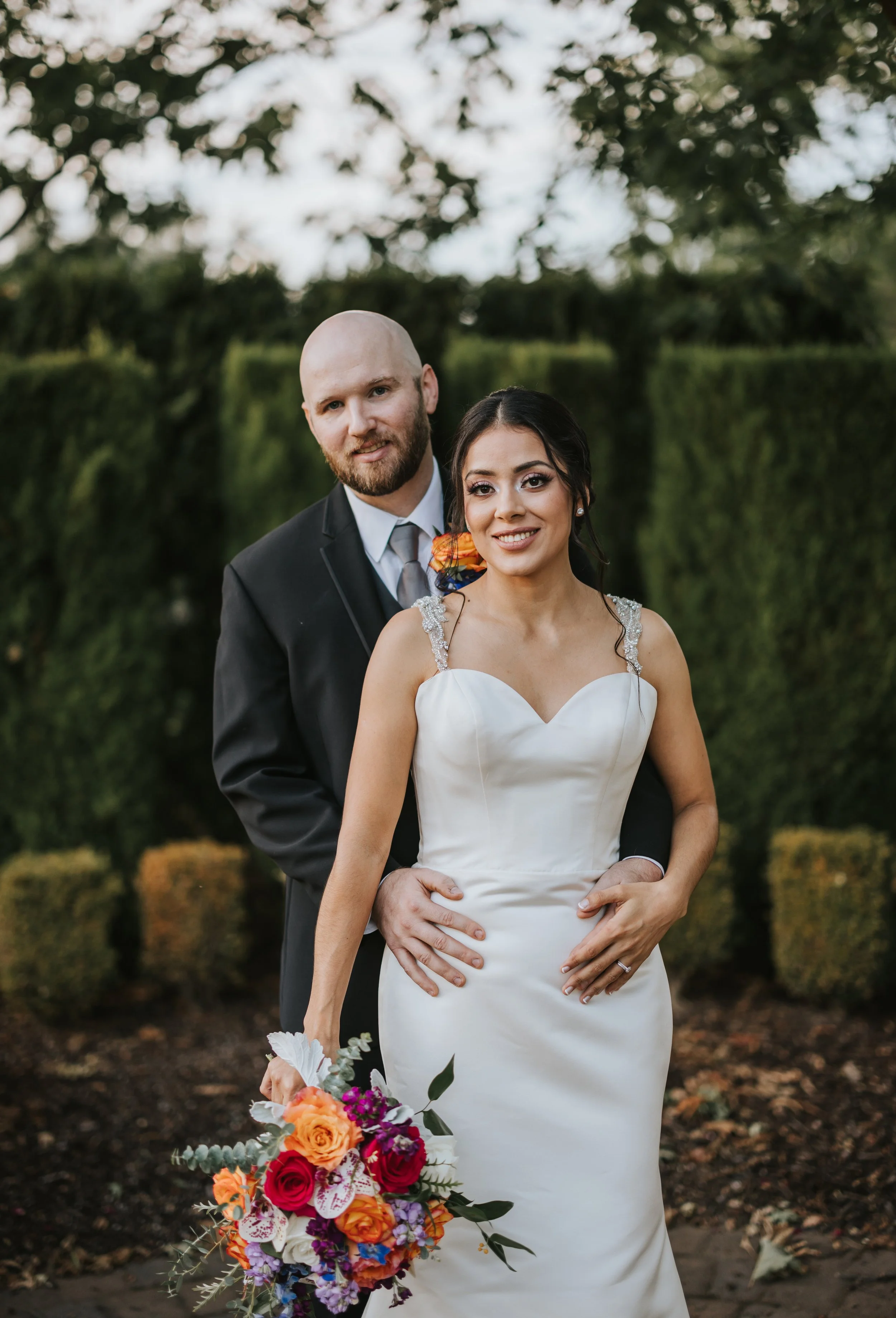 A newlywed couple standing outdoors, the bride in a white gown with beaded straps holding a colorful bouquet, and the groom in a black suit with a light tie, with green foliage in the background.