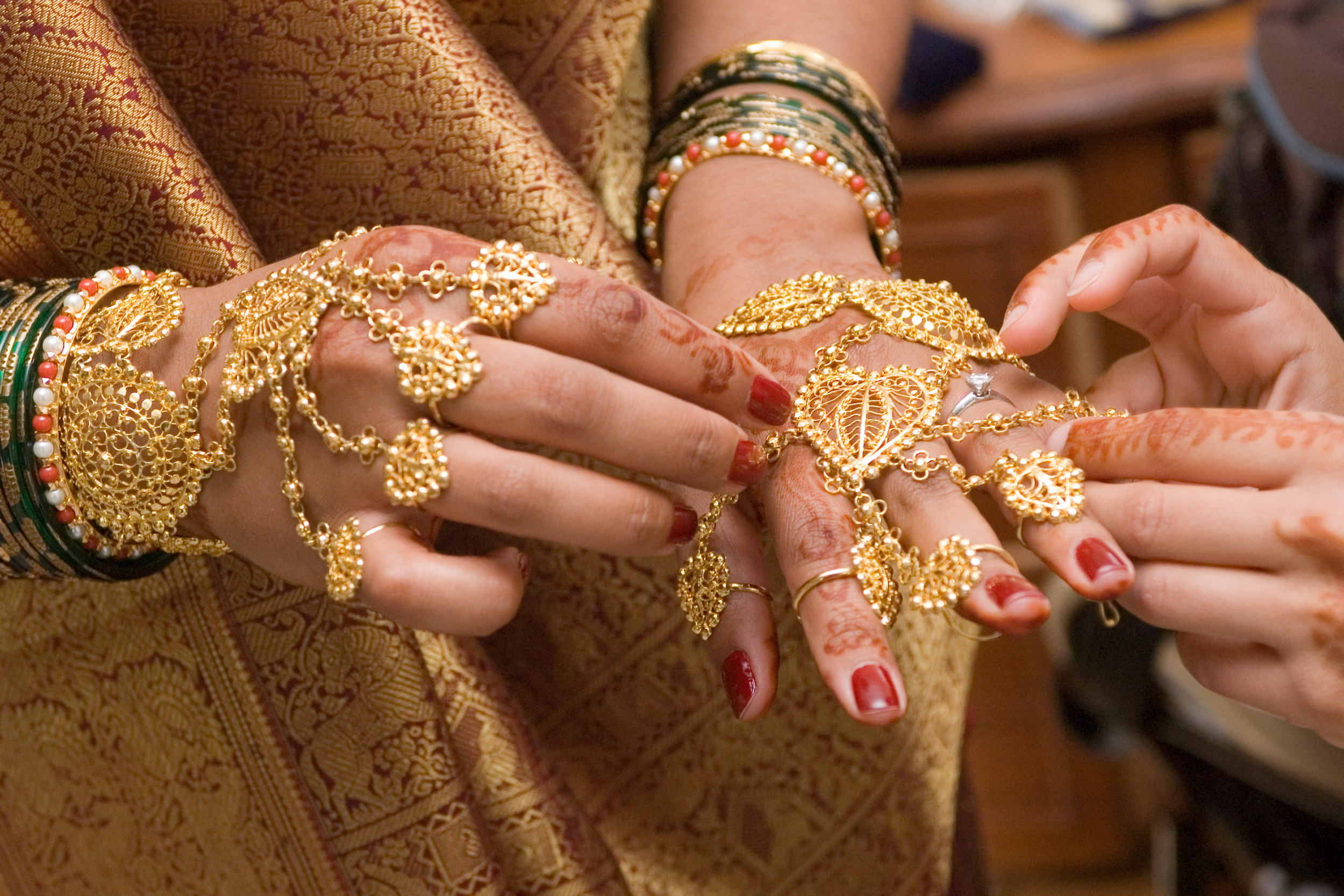 Close-up of hands adorned with intricate gold jewelry, including bracelets, rings, and chains, with henna designs on fingers. The person is wearing traditional clothing with a detailed gold and red pattern.
