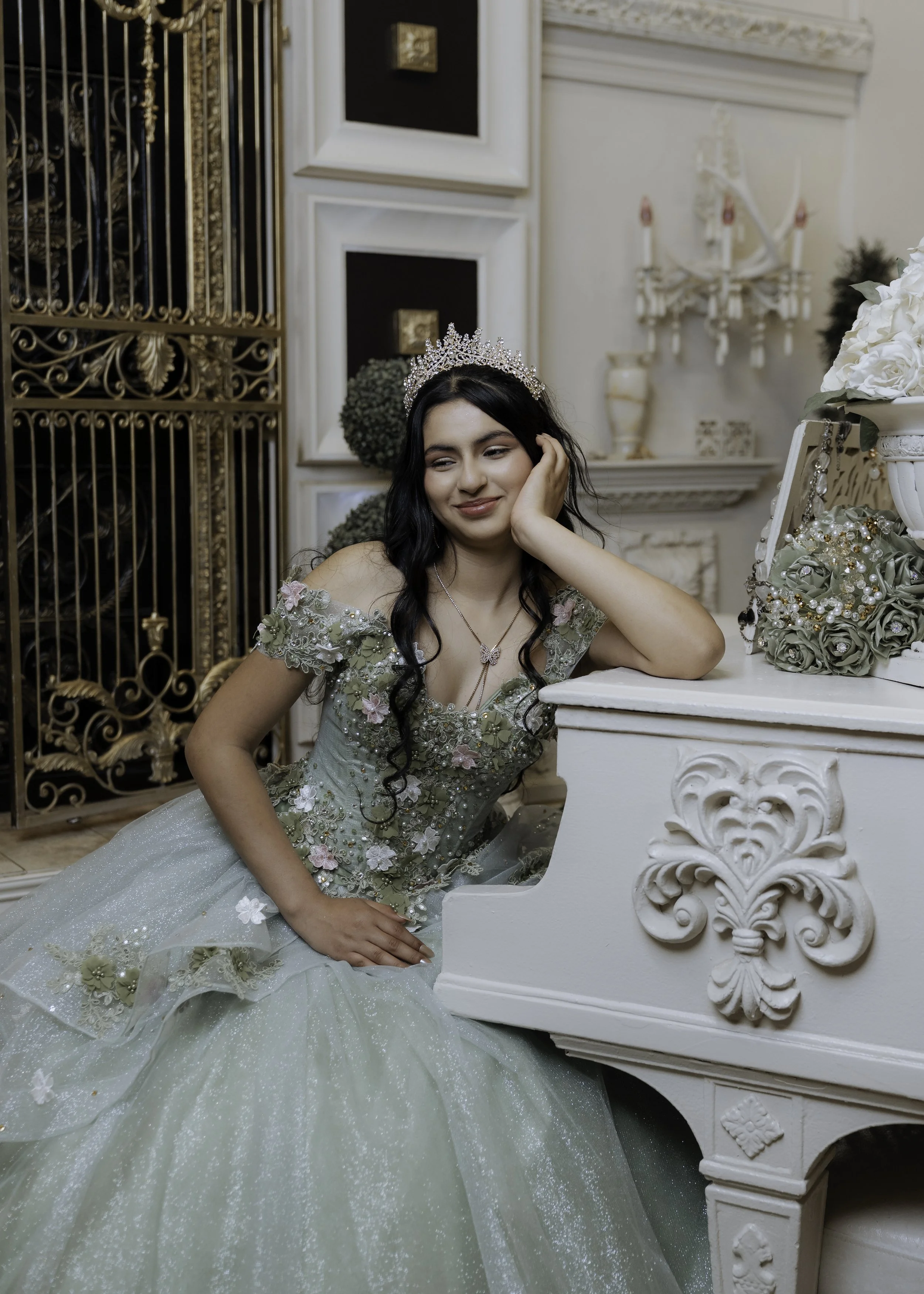 A young woman in a pastel green ball gown with floral embroidery, wearing a crown, sitting at a white ornate piano, in a decorated room with plants, flowers, a chandelier, and decorative wall framing.