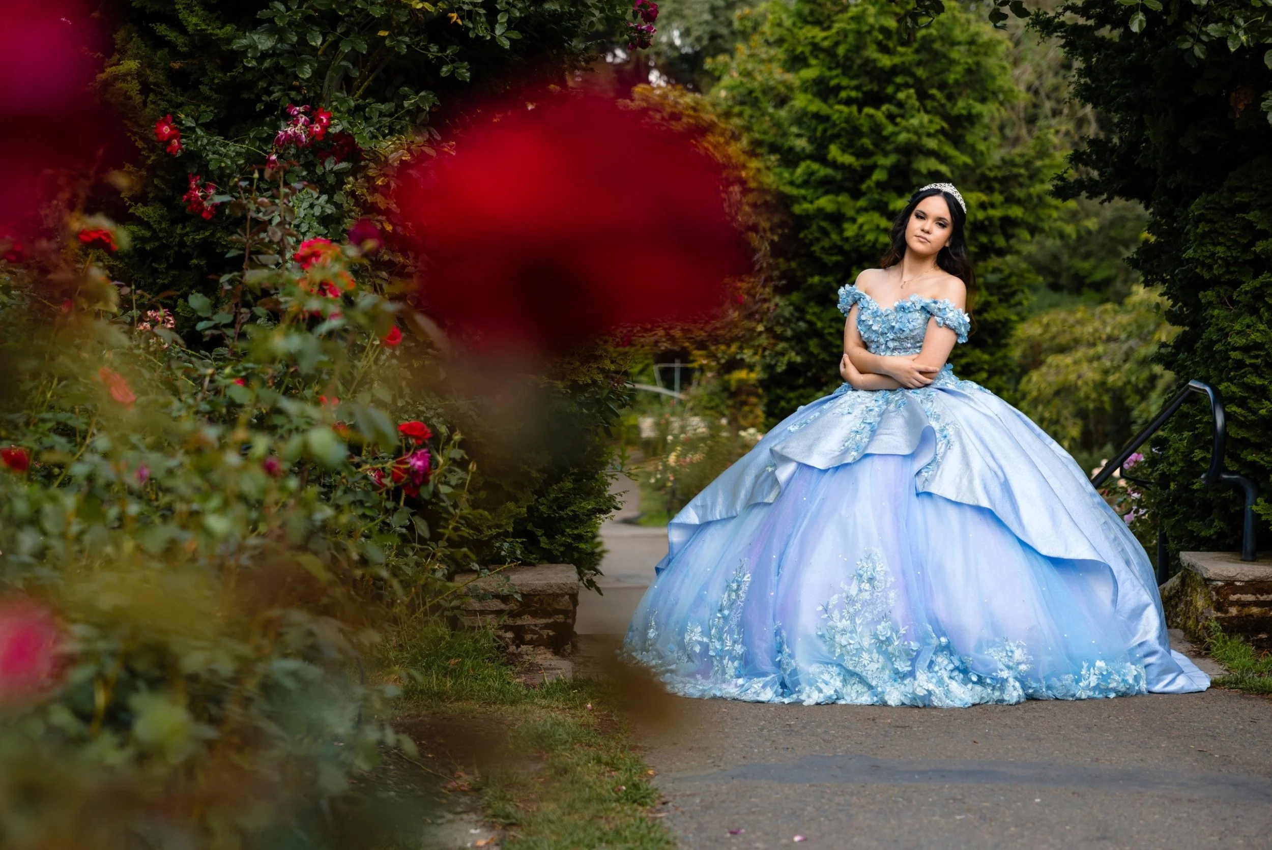 A young woman in a sky blue princess ball gown with floral details, standing outdoors on a stone pathway surrounded by lush green trees and bushes, with pink and red flowers in the foreground.
