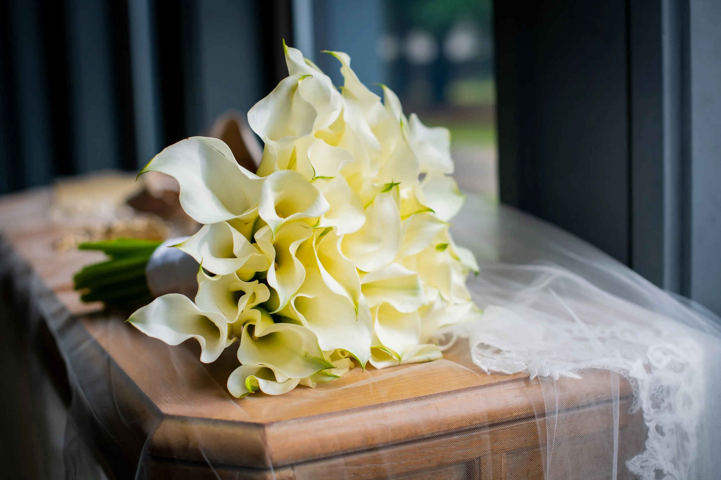 Cream-colored calla lilies bouquet placed on a wooden table near a window with dark curtians.