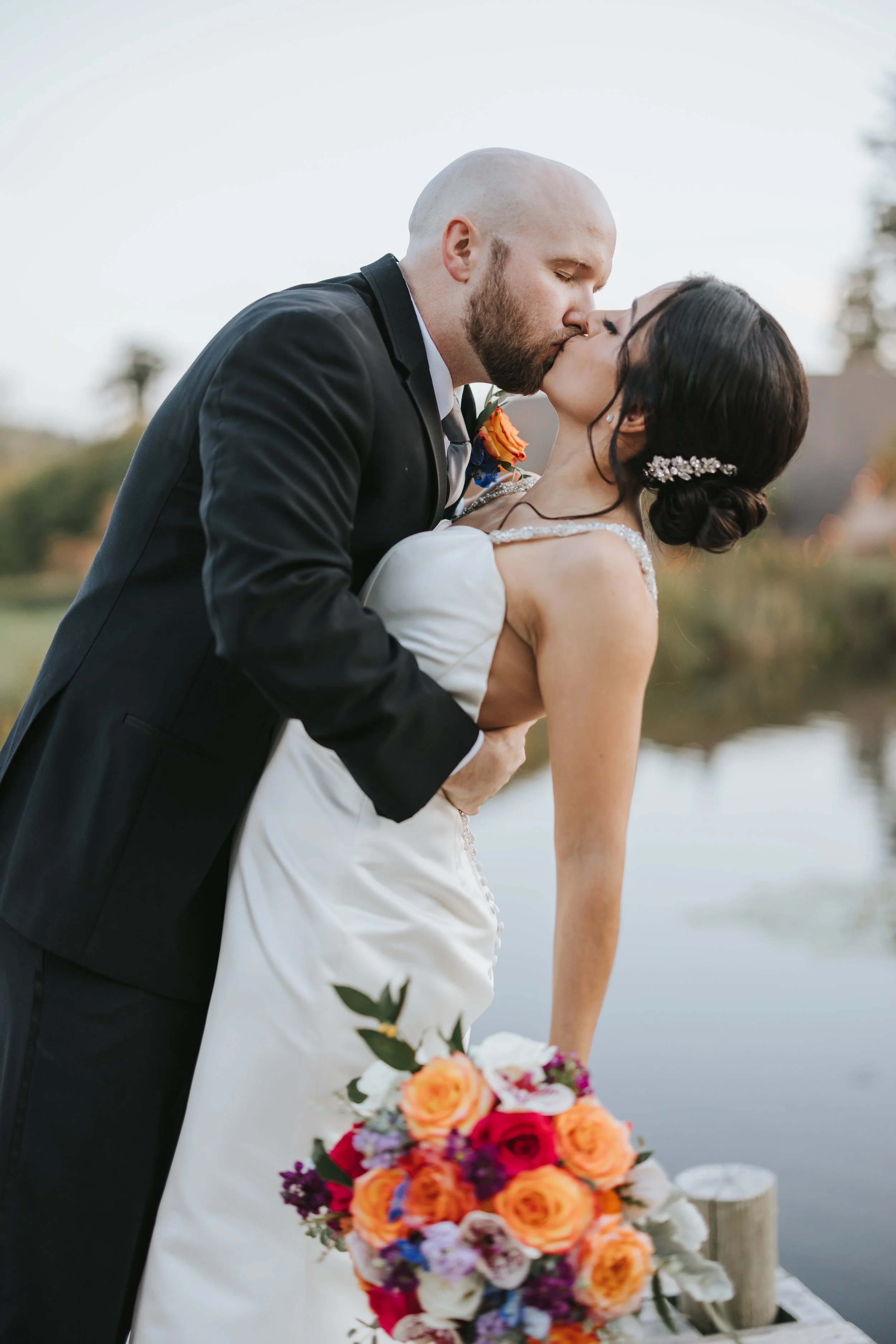 A newlywed couple sharing a kiss outdoors near a body of water, the bride holding a vibrant bouquet of orange, pink, and purple flowers.