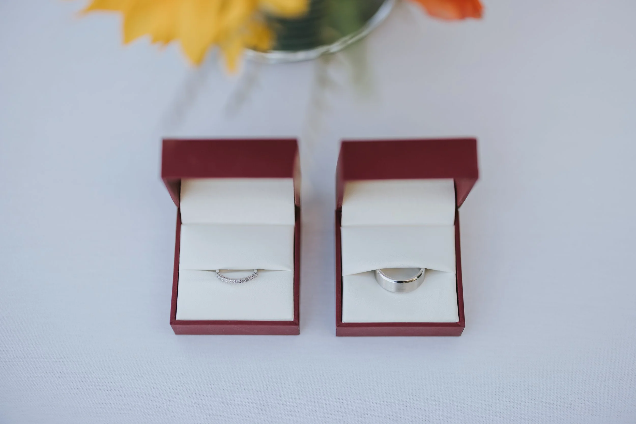 Two red jewelry boxes, one with a diamond ring and the other with a silver wedding band, placed on a white surface with a blurred floral arrangement in the background.