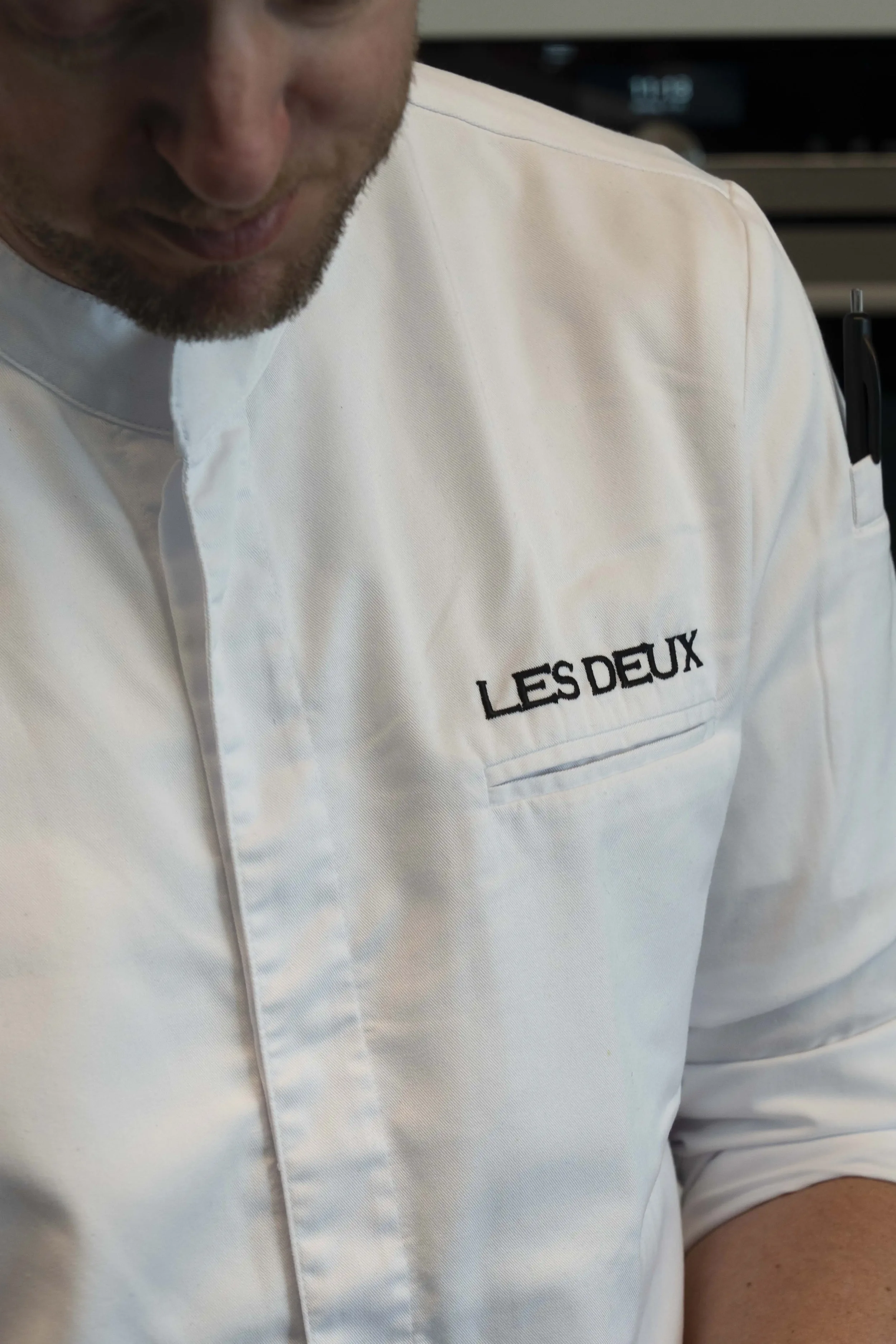 Detail van de koksvest met het logo van Restaurant Les Deux tijdens de bereiding van een gerecht.