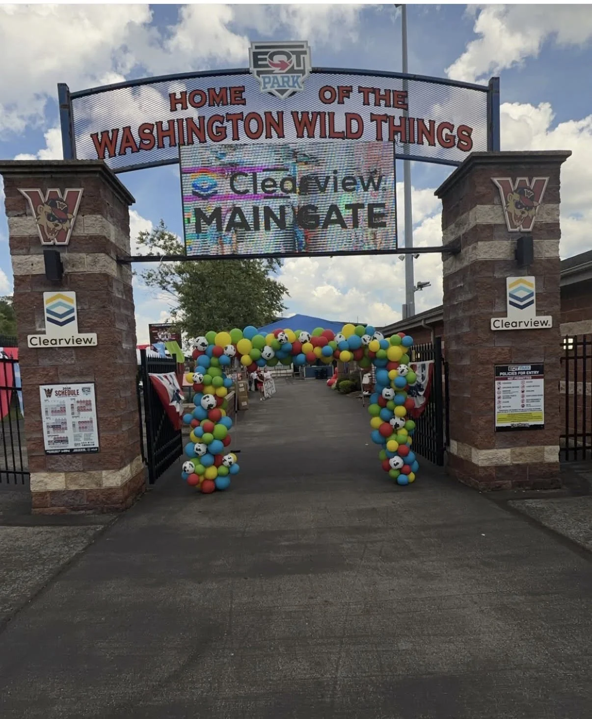 Entrance to Washington Wild Things baseball game or event with colorful balloon arch decorated with soccer balls, under a sign reading "Home of the Washington Wild Things," and a large digital sign displaying advertisements, on a partly cloudy day.