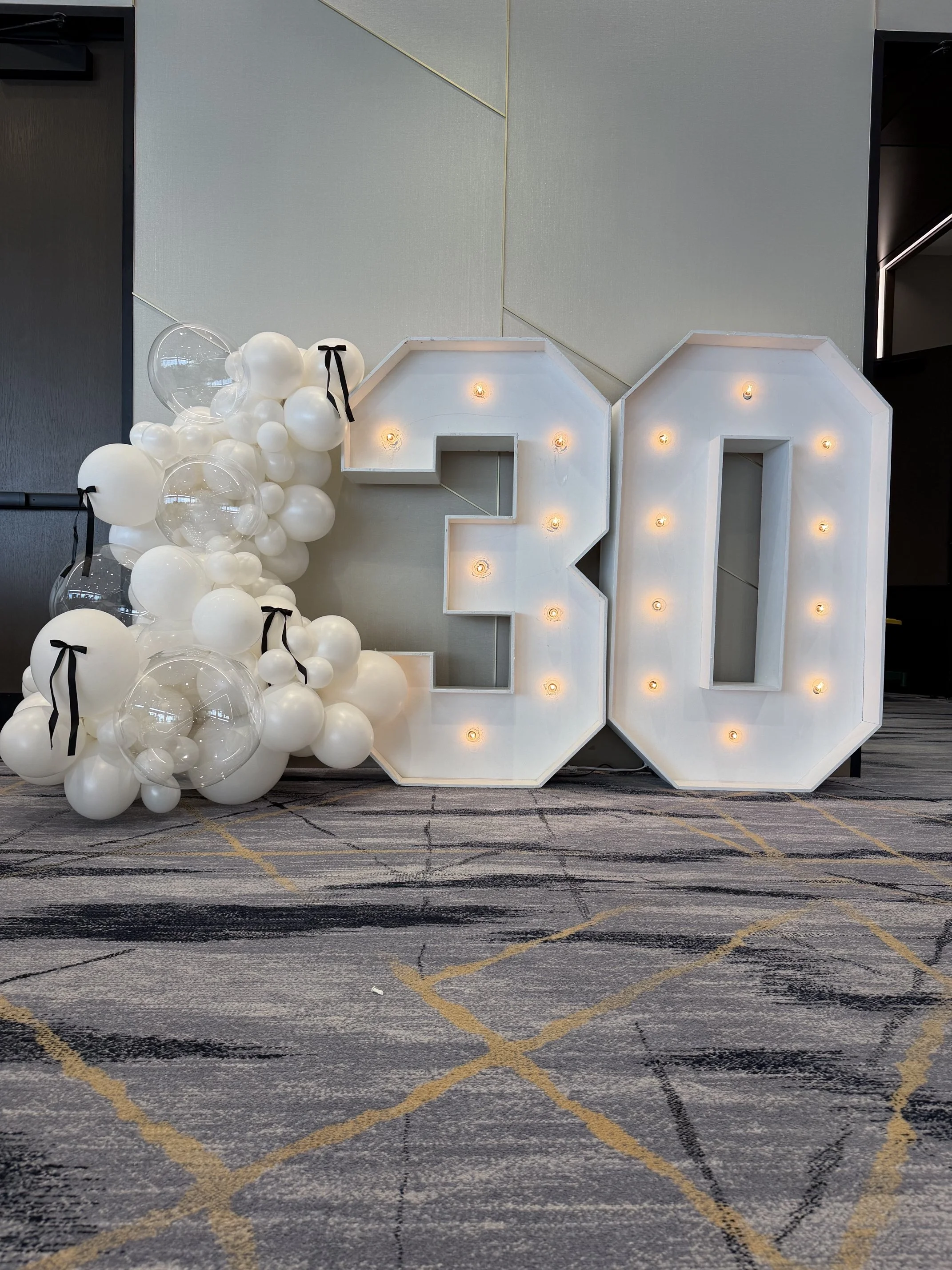 Decorative display featuring illuminated white marquee letters 'F' and 'A' with white balloons on the left side, set on a patterned carpeted floor area.