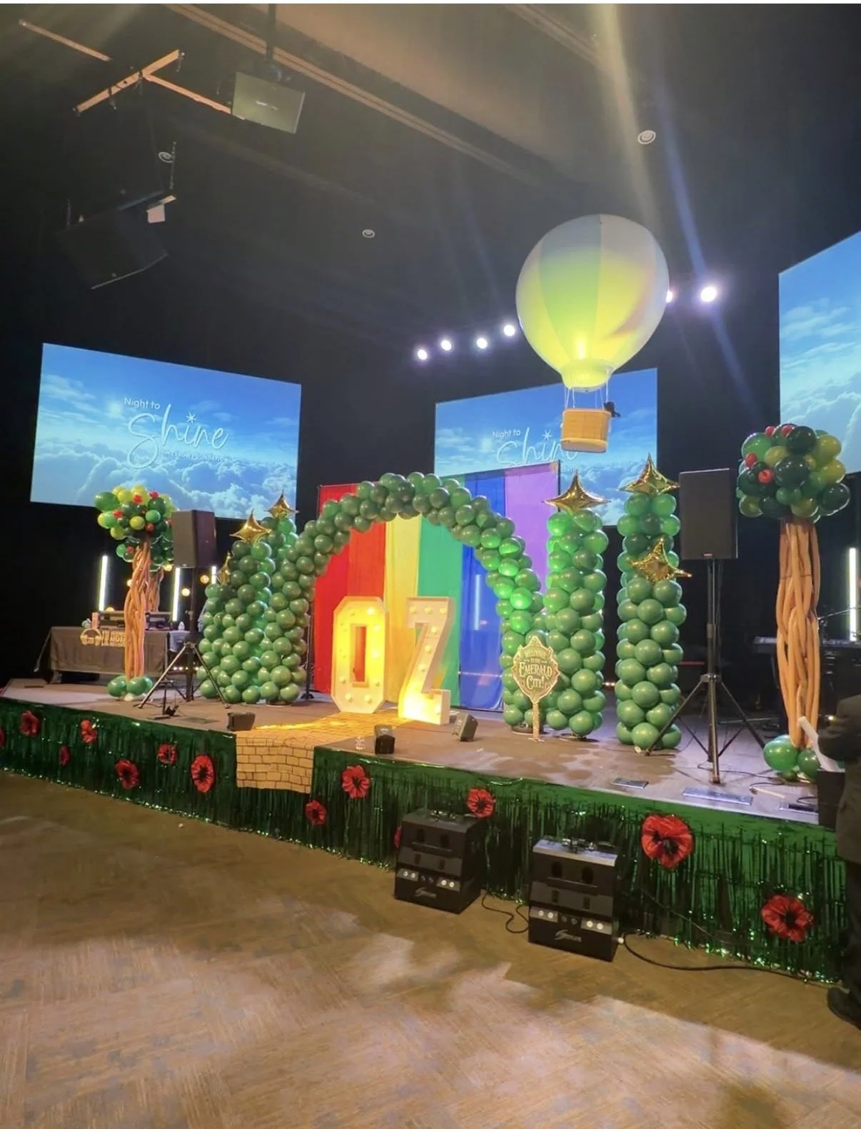 Celebration stage decorated with green balloon trees, colorful backdrop, large illuminated 'OZ' letters, a hot air balloon prop hanging from the ceiling, and a banner with the phrase 'Night to Shine.'