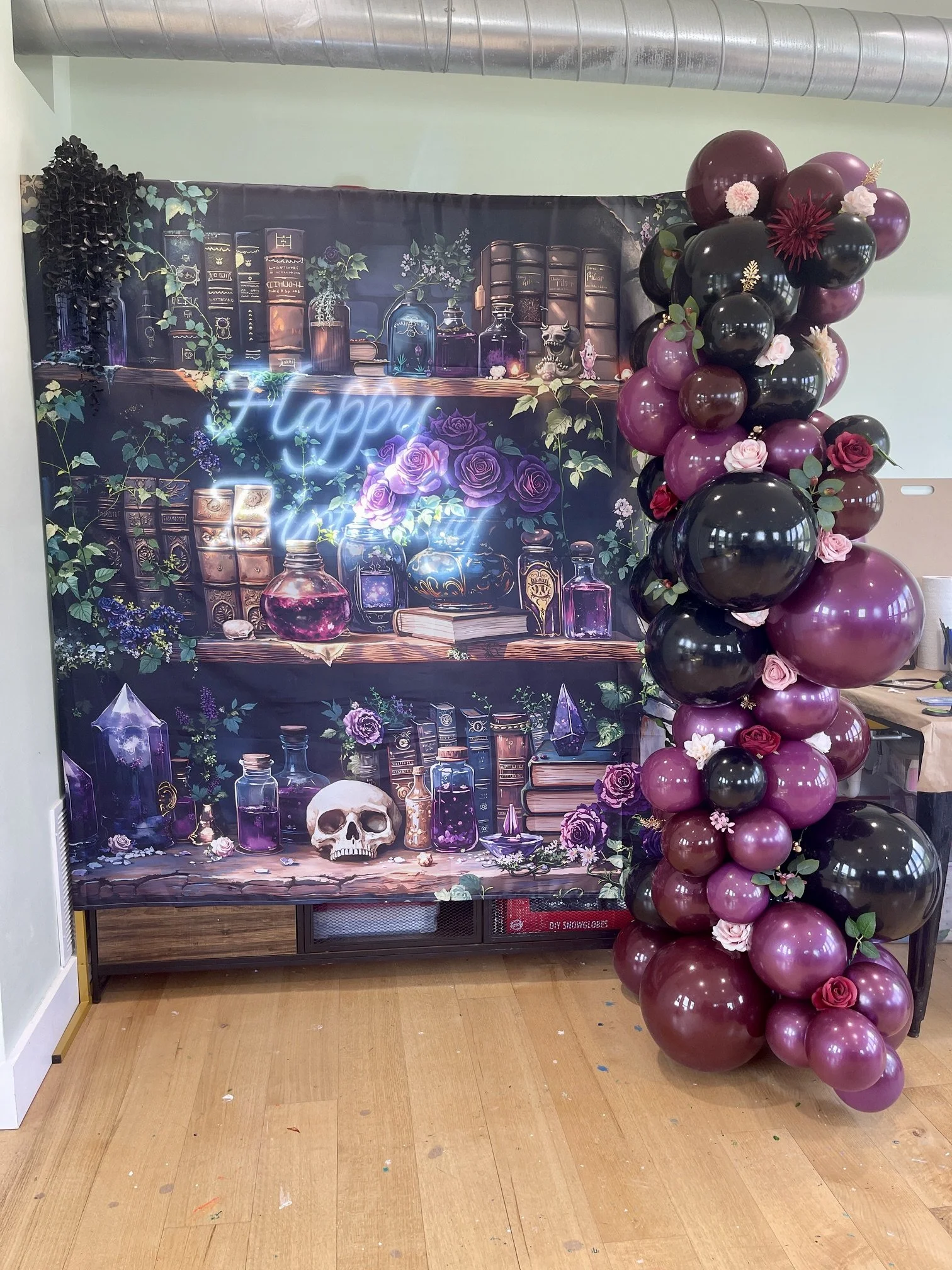 A decorative backdrop with a dark theme featuring books, potion bottles, skulls, and purple roses, decorated with balloons in shades of purple, black, and pink, with some flowers and leaves attached.