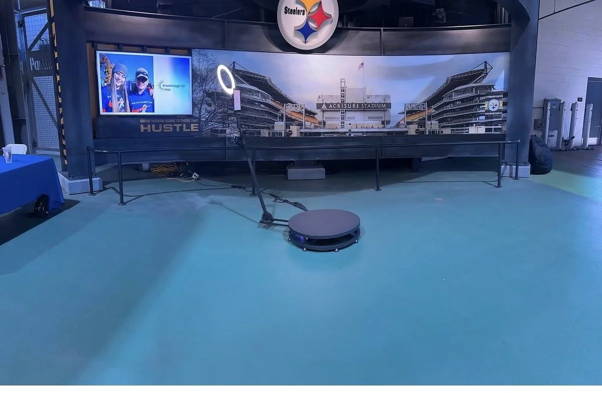 A 360 Photo Booth in front of a Pittsburgh Steelers stadium backdrop at Acrisure Stadium, with a large Steelers logo overhead and TV screen to the side.