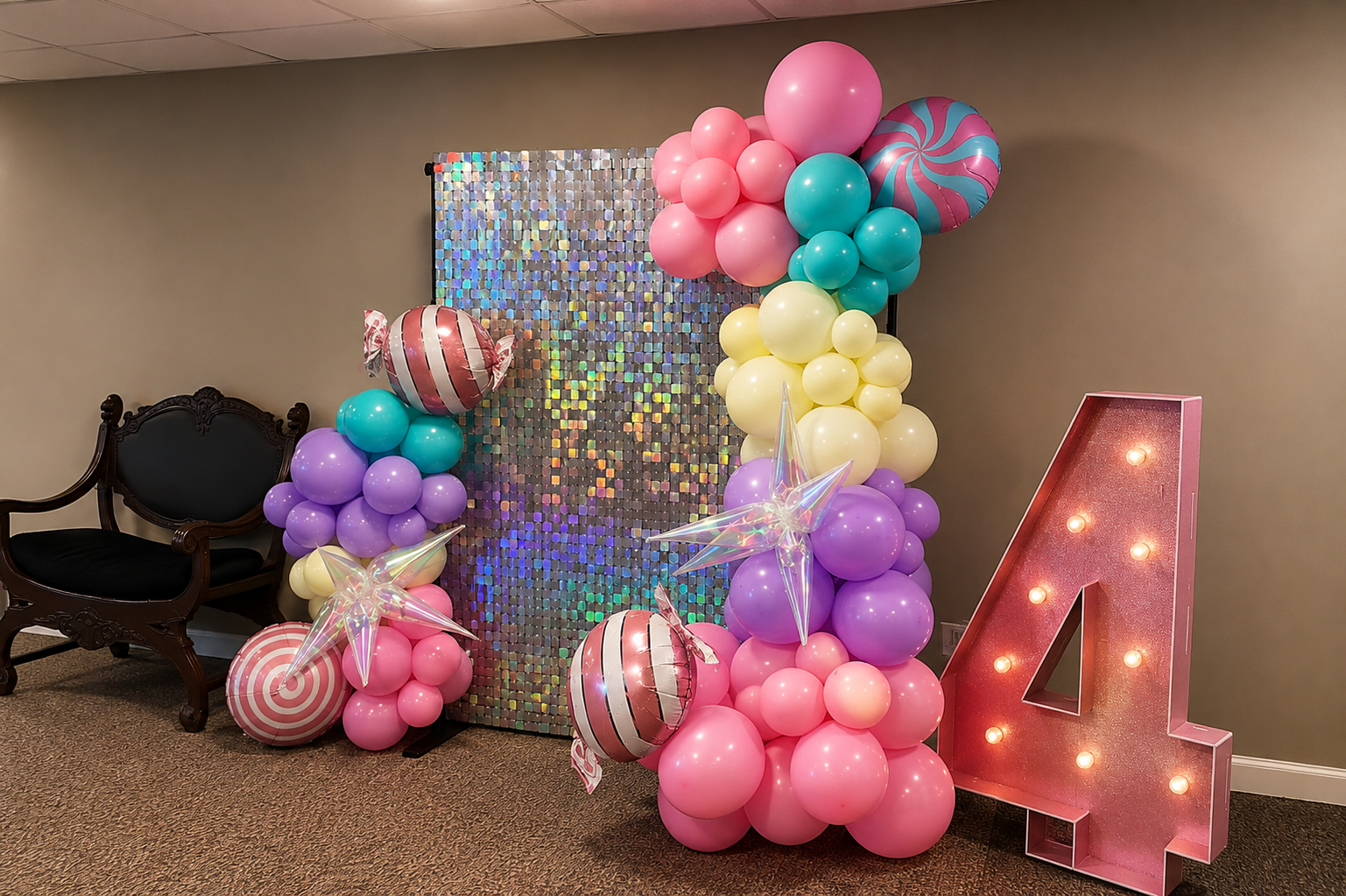 Colorful balloon decoration with pink, purple, yellow, turquoise, and patterned balloons surrounding a shiny, iridescent backdrop and a large pink letter 'A' with lights.
