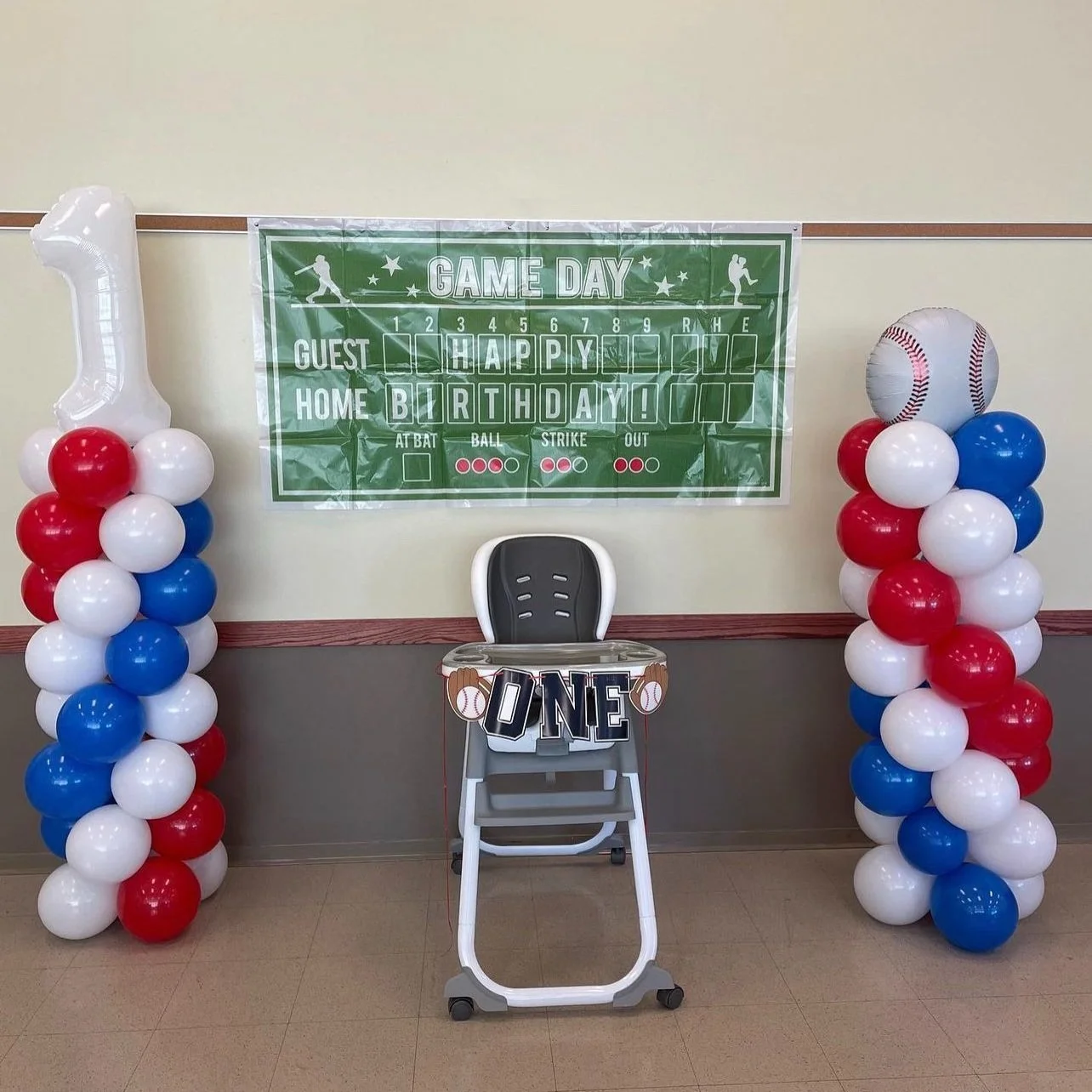 Decorations for a baseball-themed birthday celebration with red, white, and blue balloons on either side of a high chair, a baseball balloon on the right, and a green game day poster with a scoreboard highlighting 'Happy Birthday' and the number one.
