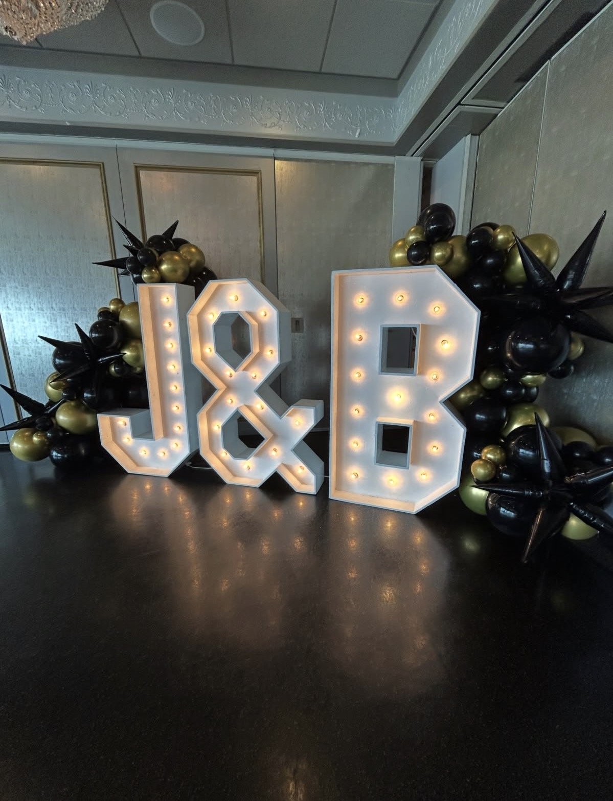 Decorative illuminated letters 'J' and '8' surrounded by black and gold balloons with pointed black balloon spikes, set against light-colored ornate wall panels.