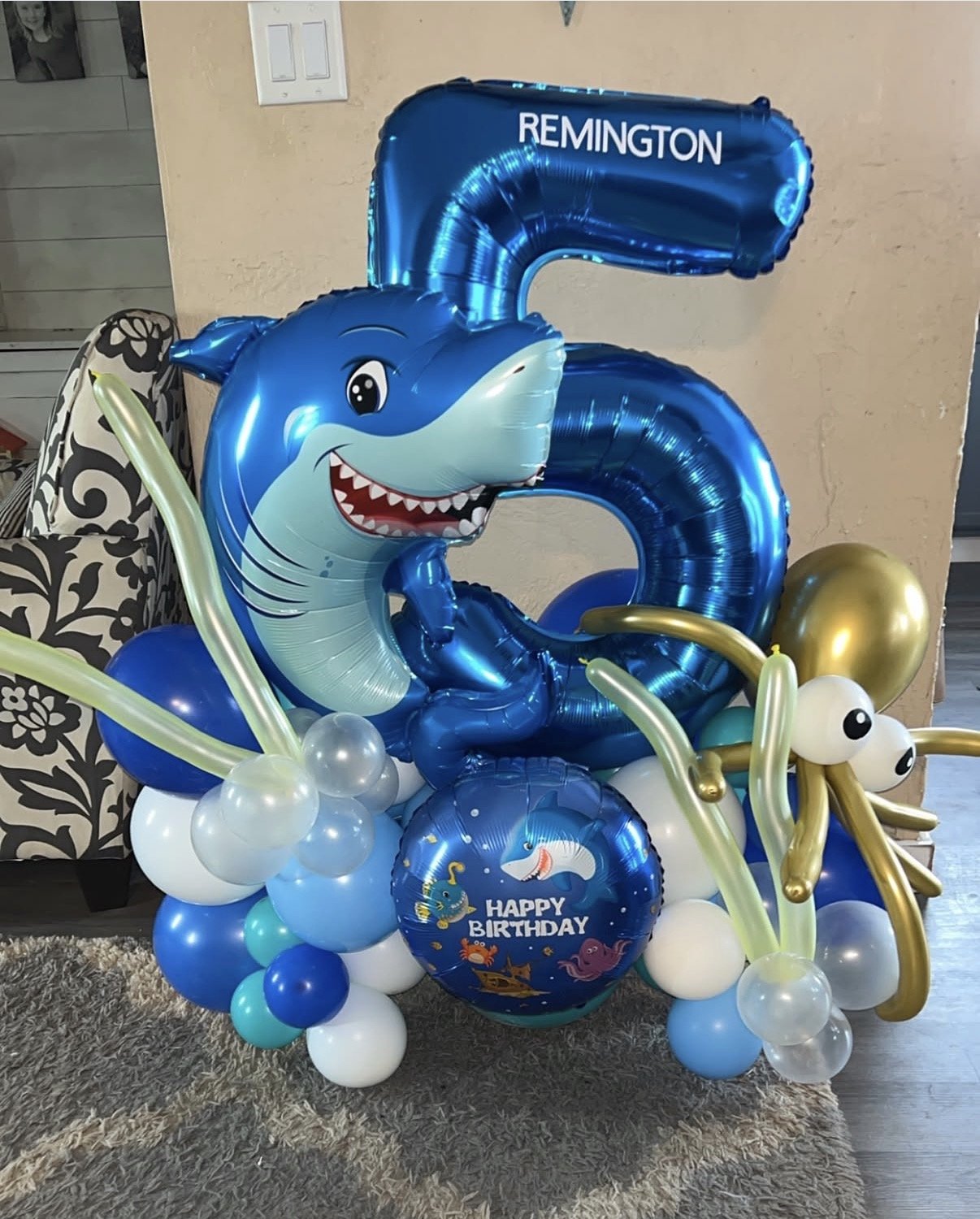 Blue shark balloon with a big smiling face, surrounded by white, blue, and gold balloons, and a blue balloon with a 'Happy Birthday' message and ocean-themed illustrations.