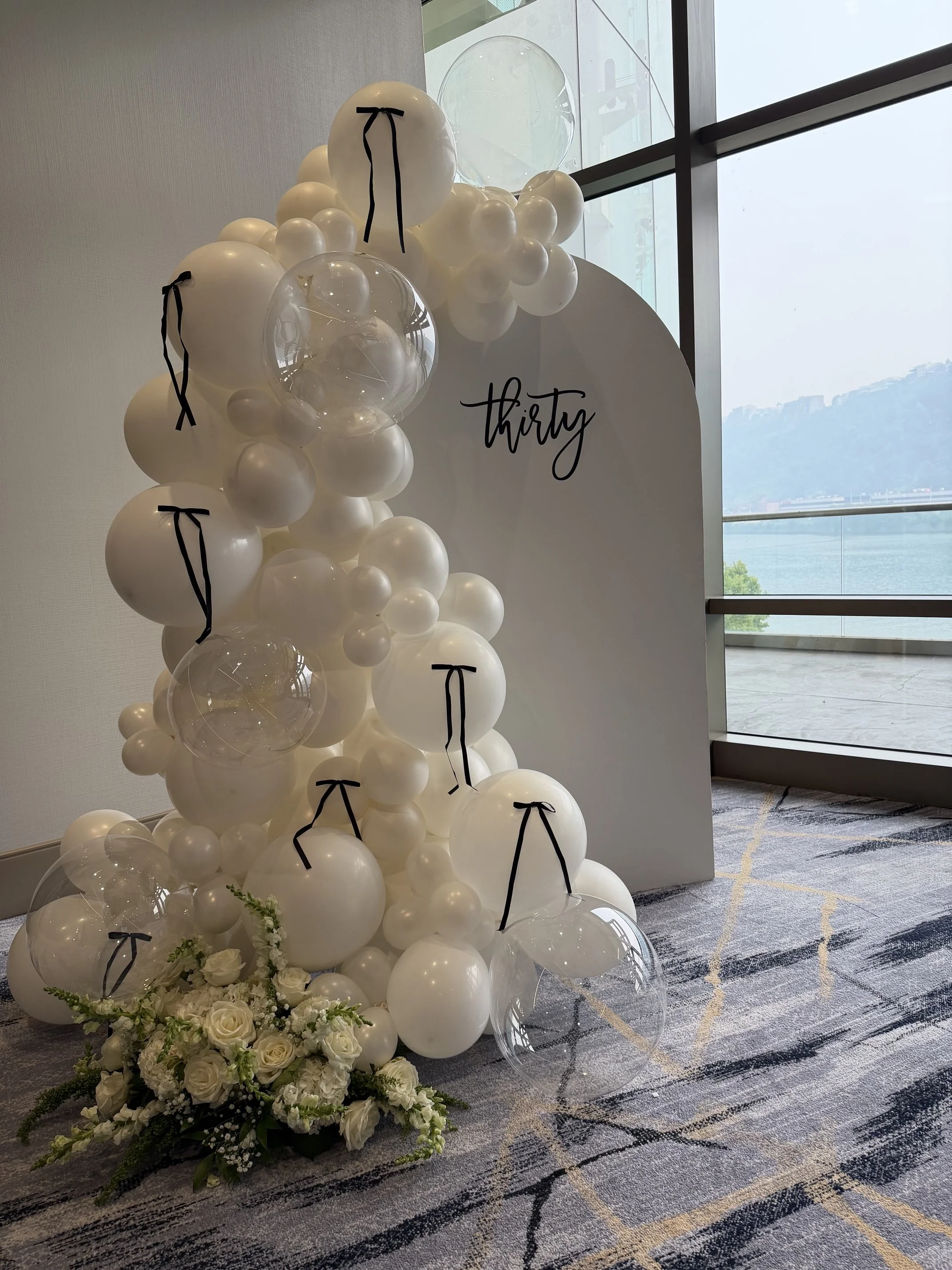 Decorative balloon display with white balloons and black ribbon accents, next to a floral arrangement of white roses and greenery, with a white circular sign that reads 'thirty' in black cursive font, positioned indoors near large windows overlooking